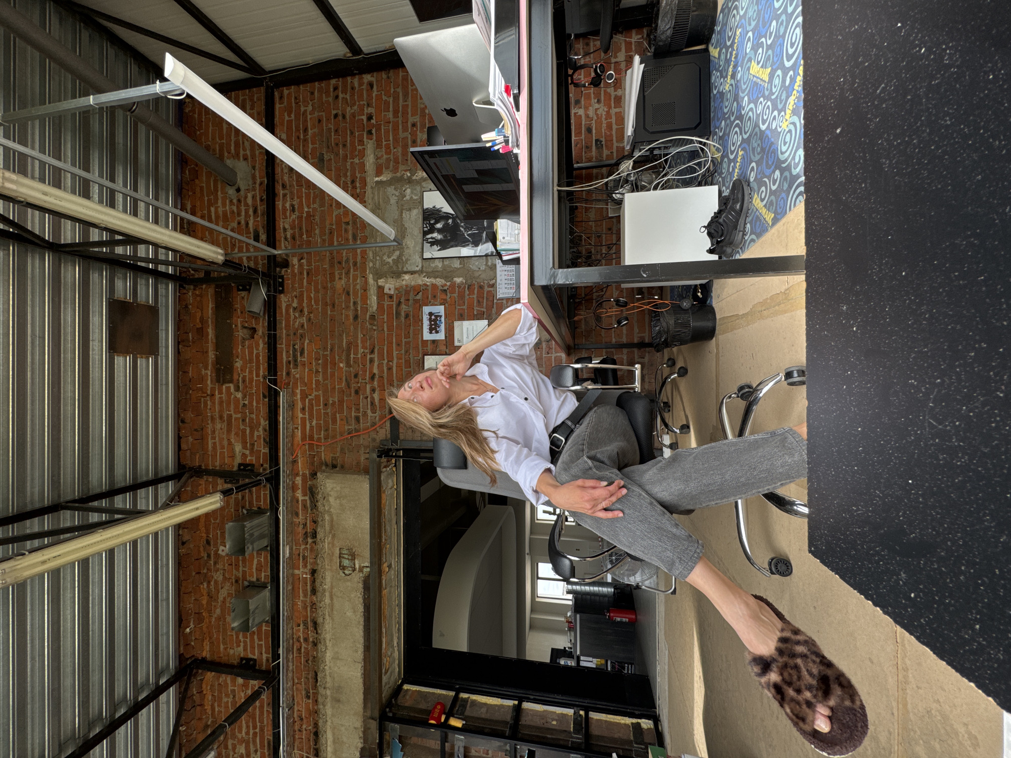 A woman with long blonde hair, wearing a white button-down shirt and grey jeans, sits in a black office chair. Her legs are crossed, and she is wearing leopard-print slippers. She rests her chin on her hand, looking upwards. The office has exposed brick walls and a metal ceiling with fluorescent lights. A desk with computers and office supplies is positioned to her right. In the background, an open doorway reveals a glimpse of another room. The setting appears to be an office space, possibly in an industrial or loft-style building, located in the city of Chaikovsky, Russia.