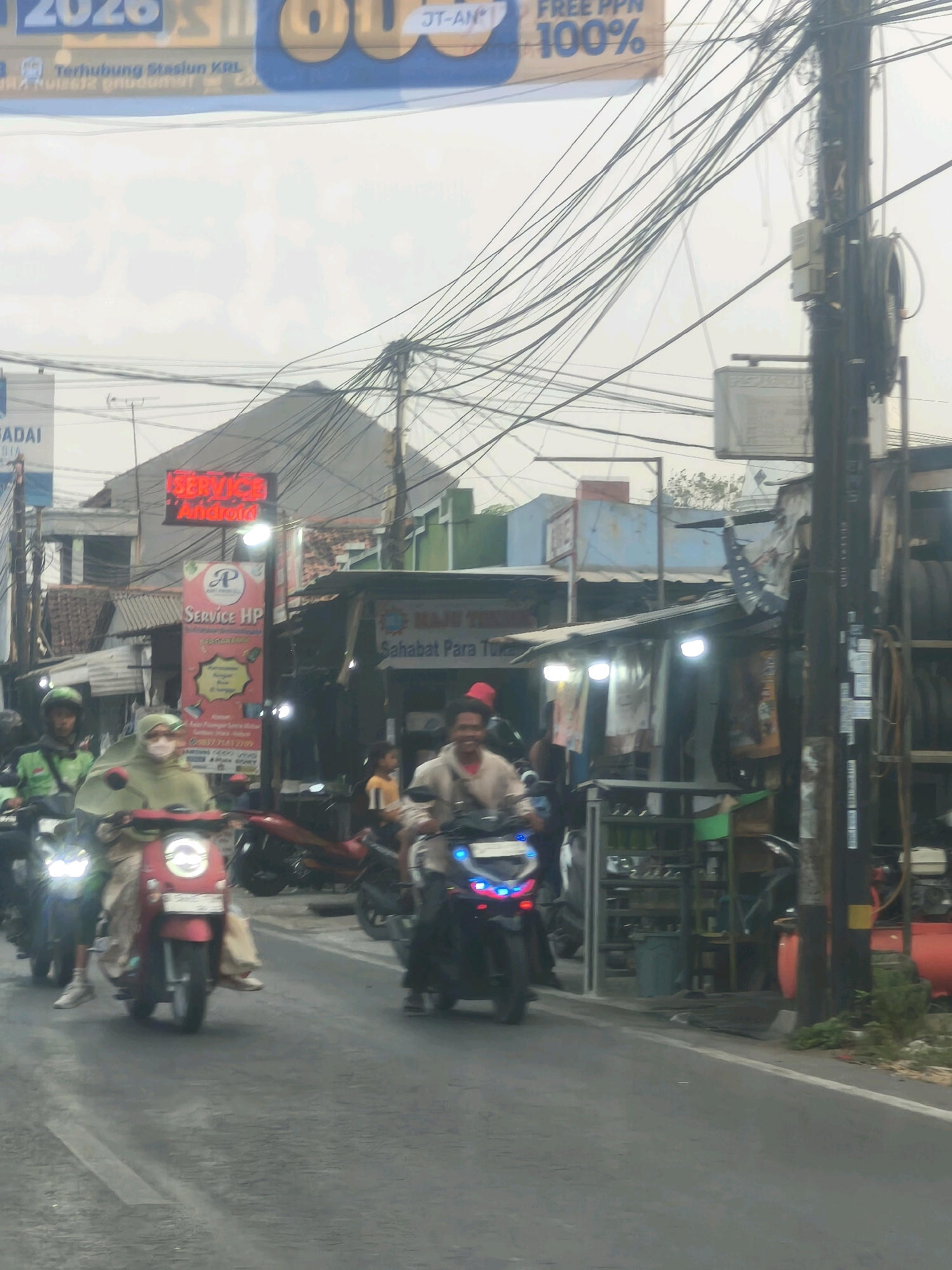 In the city of Satriamekar, Indonesia, a street scene unfolds with multiple motorcycles and their riders navigating a paved road under a cloudy sky. The foreground is dominated by a red scooter with a rider wearing a green hijab and a patterned fabric draped over their shoulders, and another motorcycle on the left with a rider in a green jacket and helmet. To the right, a motorcycle with blue flashing lights is ridden by a person wearing a light-colored shirt and a red head covering. Behind the riders, a row of shops line the street. One shop prominently displays a red neon sign that reads "SERVICE Android." Another sign, vertically oriented and featuring a pink background with a logo, advertises "Service HP." Further down the street, an open-fronted shop showcases various items, including what appear to be garments hanging on display and shelves stocked with goods. Overhead, a dense network of electrical wires crisscrosses the sky, suspended from utility poles, one of which is visible on the right side of the image. The environment appears to be an urban or semi-urban commercial area.