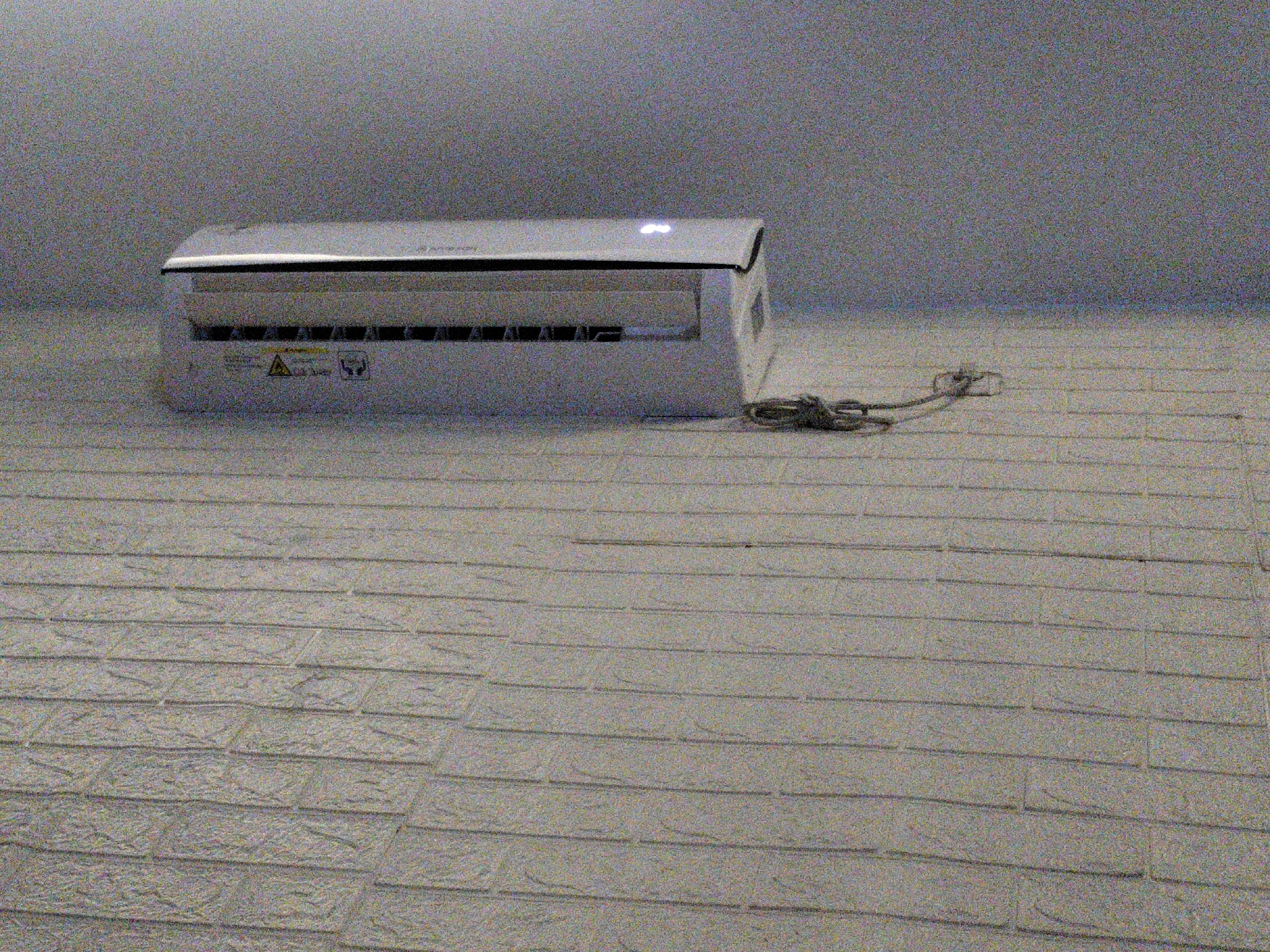 An indoor, eye-level, medium shot shows a white air conditioning unit mounted on a white, brick-patterned wall. The air conditioner is on the left side of the frame, and its front vent is open, revealing louvers. A small digital display on the top of the air conditioner shows a glowing blue '88'. To the right of the air conditioner, a black power cord is coiled on the floor and appears to be plugged into a discreet outlet. The lighting is dim, with a slight graininess affecting the image quality. The environment appears to be a domestic or commercial space, possibly a bedroom or living area, as suggested by the presence of the air conditioner. The wall has a textured, brick-like pattern. The overall scene is static and devoid of human presence.