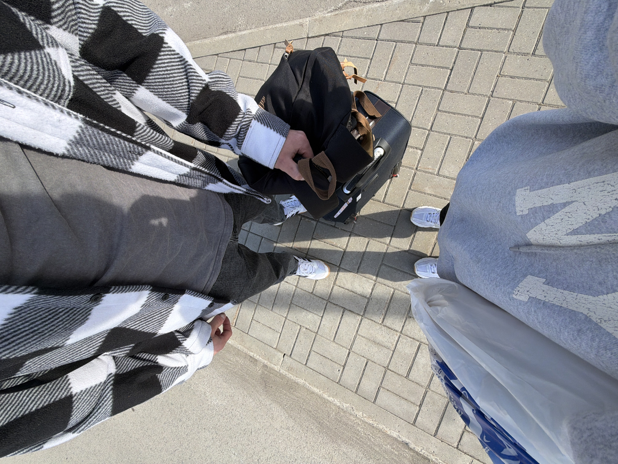 The image, taken from a high-angle perspective, shows two individuals standing on a brick-paved surface. On the left, a person is wearing a black and white plaid flannel shirt, a grey top, and dark trousers, holding a black bag on top of a suitcase. The suitcase is on wheels and has a retractable handle. They are wearing white Adidas sneakers. On the right, another person is wearing a grey hoodie and white sneakers. They are holding a large white plastic bag. The background is a brick-paved surface, partially with pavement and partially with the street. Given the absence of specific contextual details, the location cannot be definitively placed in Boldino, Russia.