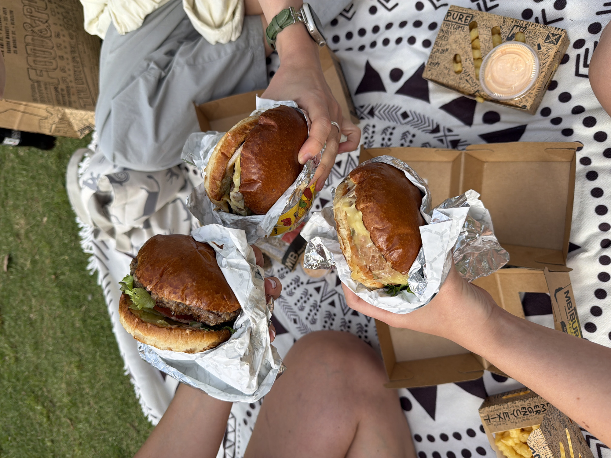The image depicts three individuals enjoying a picnic, seemingly in an outdoor setting, possibly a park, given the presence of grass visible in the lower left. Each person is holding a hamburger wrapped in foil. The burgers have visible contents like lettuce, tomatoes, and cheese. Boxes of French fries are present with a container of sauce. There are boxes from a food establishment visible, one reading "PURE" and another with Japanese text. The background includes a picnic blanket with a geometric pattern, and the upper section of the image shows people wearing casual attire. The scene suggests an informal, outdoor meal being shared. Though not explicitly stated, this could potentially be in a city like Tel-Aviv, Israel, assuming that's where these businesses operate.