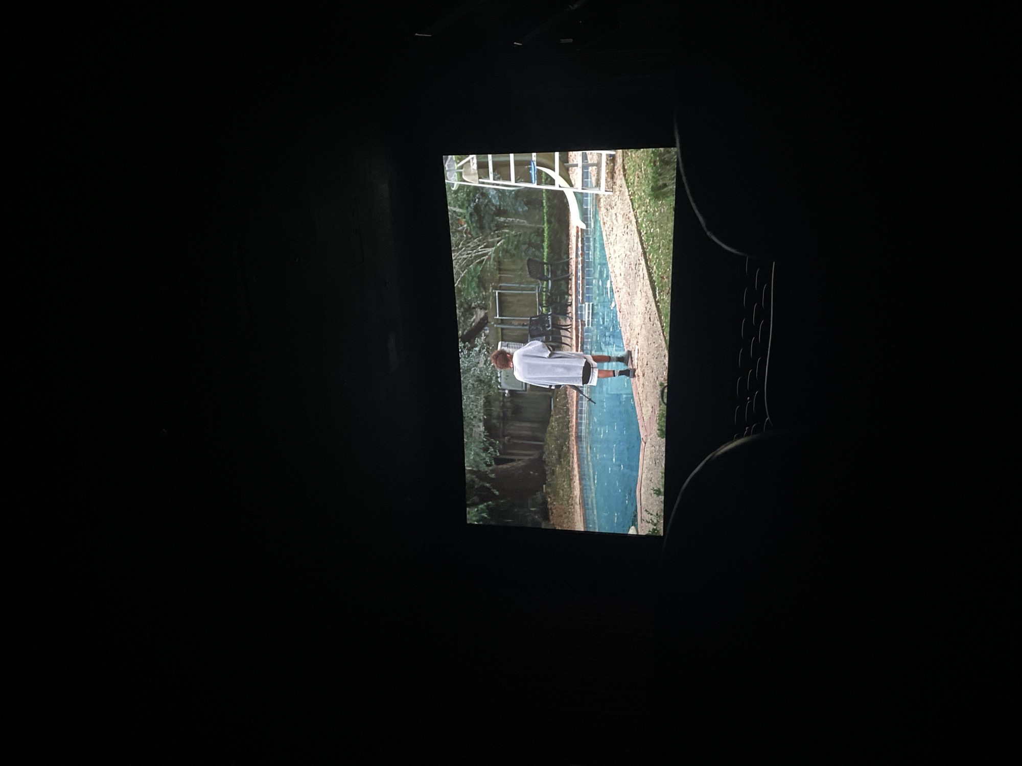 The image shows a person standing near a swimming pool, visible through what appears to be a projection screen. The person is wearing a light-colored shirt and dark shorts, and is standing on the pool deck. The pool has a slide and ladder. Behind the pool there are chairs and trees. The surroundings are very dark, as though the picture on the screen is being viewed in a dark room. The location is undeterminable.