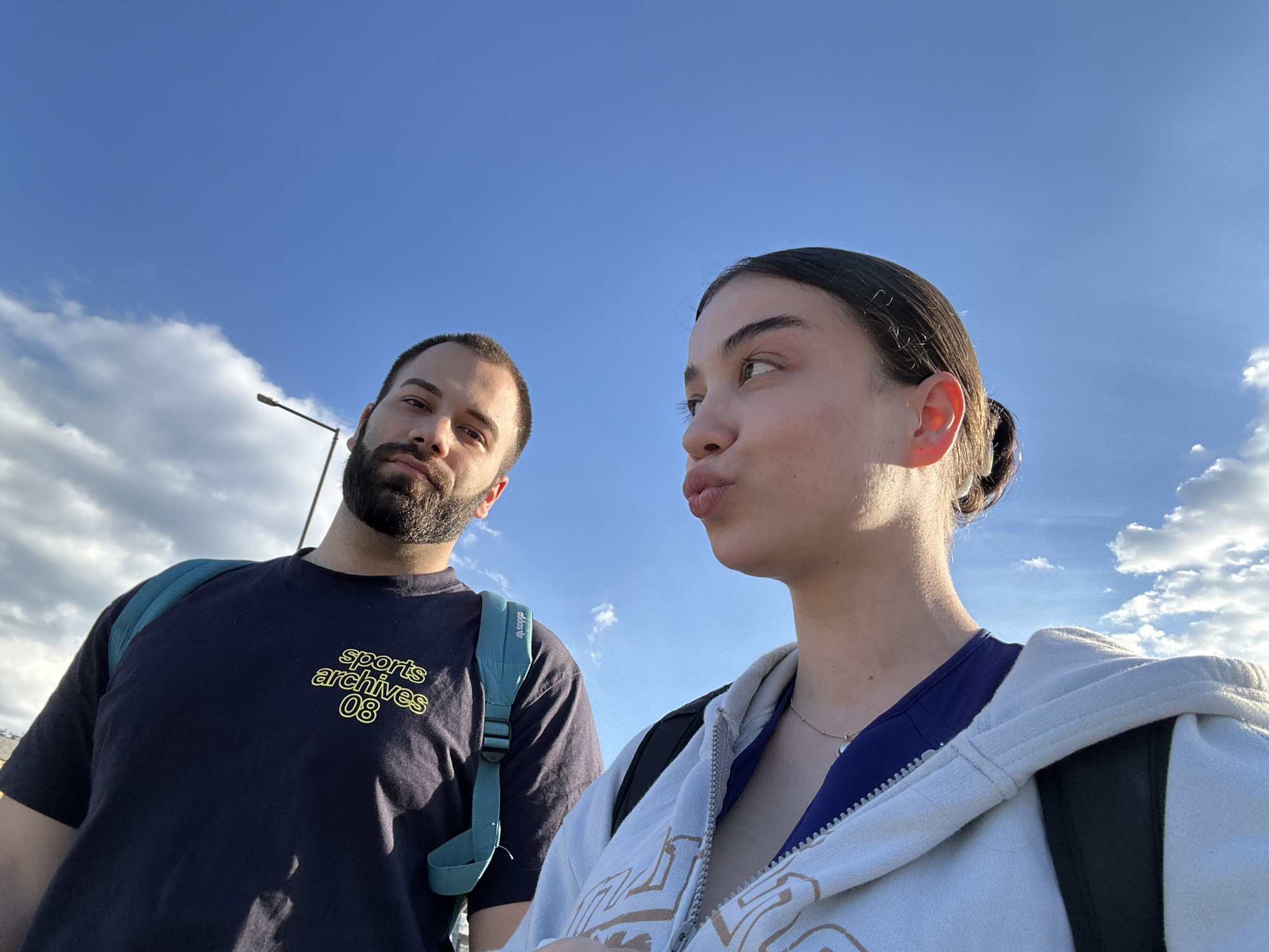 The image depicts two individuals, a man and a woman, likely taking a selfie under a bright blue sky with scattered clouds. The man, on the left, has a beard and is wearing a dark blue T-shirt with yellow text that reads "sports archives 08." He has a teal backpack on his back. The woman, on the right, has her hair pulled back and is wearing a white hooded sweatshirt over a dark blue top. She is looking to the side with pursed lips. A street light is visible in the background. The image does not provide enough information to establish the location as the city of Sofia, Bulgaria.
