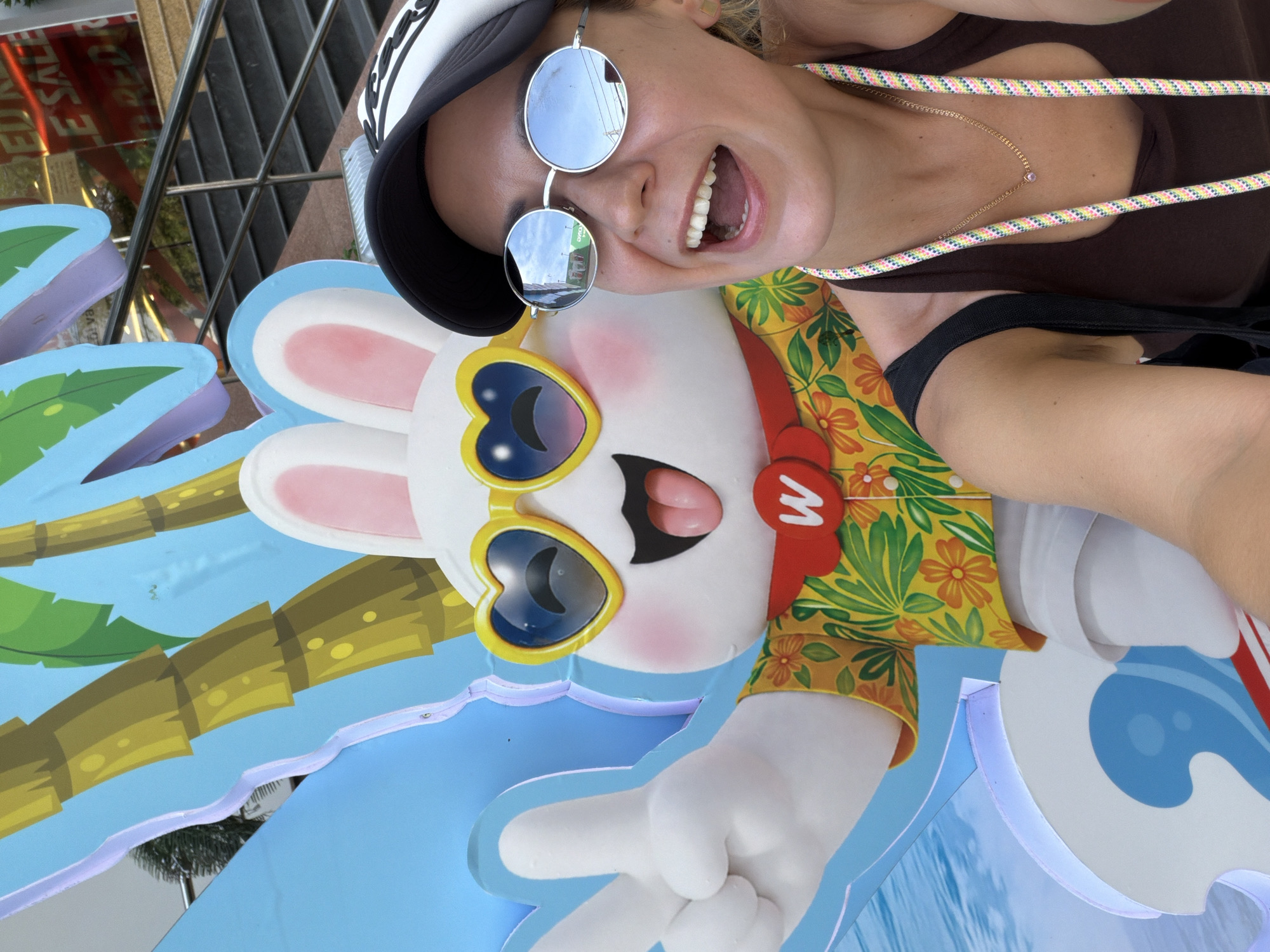 The image shows a woman taking a selfie with a large cartoon rabbit cutout. The cutout depicts a white rabbit wearing heart-shaped sunglasses and a tropical-print shirt, holding up a peace sign. The background features a bright blue sky with palm trees, suggesting a summery or tropical theme. The woman is wearing a dark tank top, a baseball cap, and sunglasses, and is smiling widely. In the background, a building with red "SALE" signage and a staircase can be seen. The scene appears to be in an outdoor urban setting, possibly a mall or a commercial area in Nha Trang, Vietnam, given the style of the signage and the architecture.