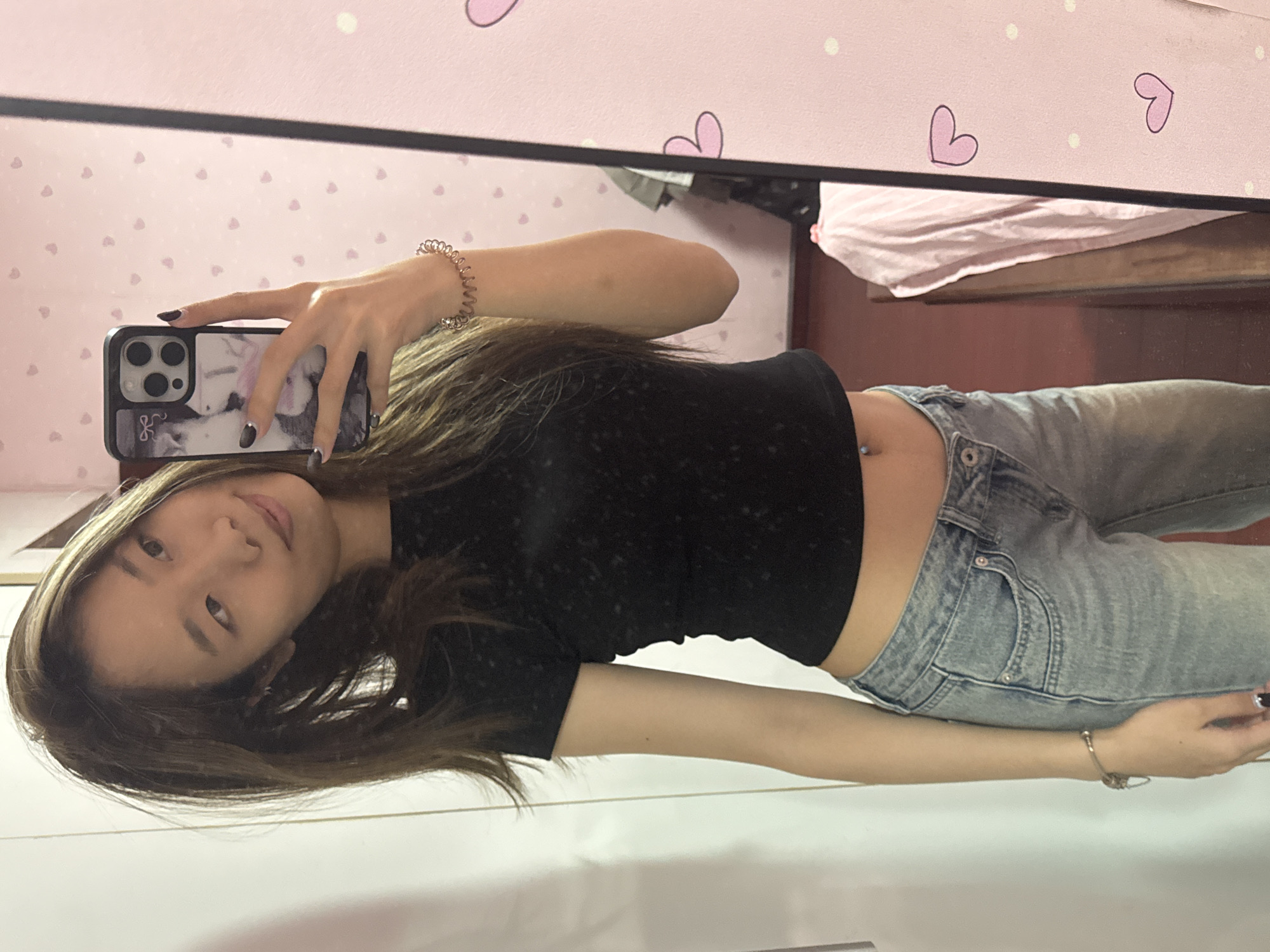 A young woman is posing in a mirror selfie. She is wearing a black crop top and light wash jeans. She has long, brown hair. She is holding a smartphone with a decorative case. The background includes a pink wall with a heart pattern, a bed with pink linens, and a portion of a wooden floor. The woman is wearing a bracelet on her left wrist and another on her right wrist. A body piercing is visible on her stomach. It is difficult to determine the precise location within Shapingba District, China, based solely on the provided image.