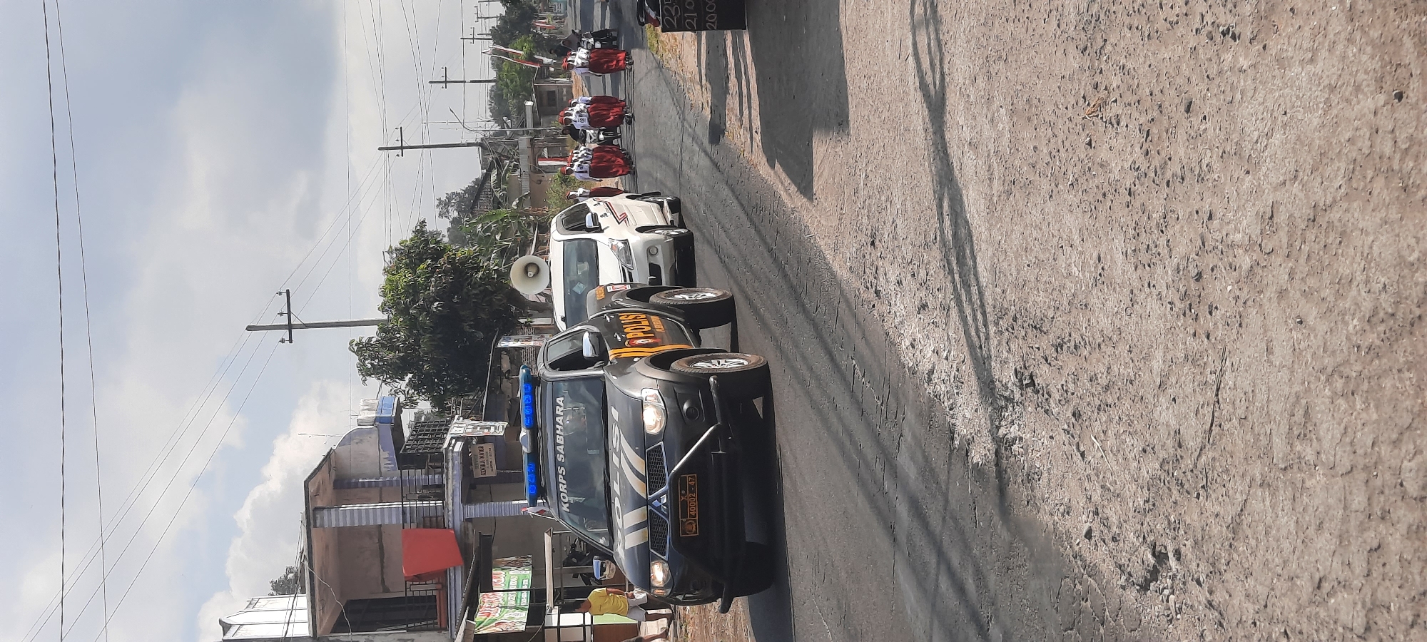 This image depicts a street scene in Jatitengah, Indonesia, possibly during a parade or procession. In the foreground, a black police pickup truck with "POLISI" and "KORPS SABHARA" written on its side is driving on a paved road. The truck has flashing blue lights on its roof and a megaphone mounted on top of a white car following it. Behind the white car, a group of individuals, appearing to be students in uniform with red skirts and white shirts, are walking in a procession. Some are carrying flags. A motorcycle with a rider is visible further down the road. Buildings and trees line the street, and utility poles with wires are present. The sky is partly cloudy. The road surface shows signs of wear and tear with cracks and uneven patches. There are also shadows cast on the road, indicating it is daytime. The overall impression is of an organized event proceeding along a moderately busy street.