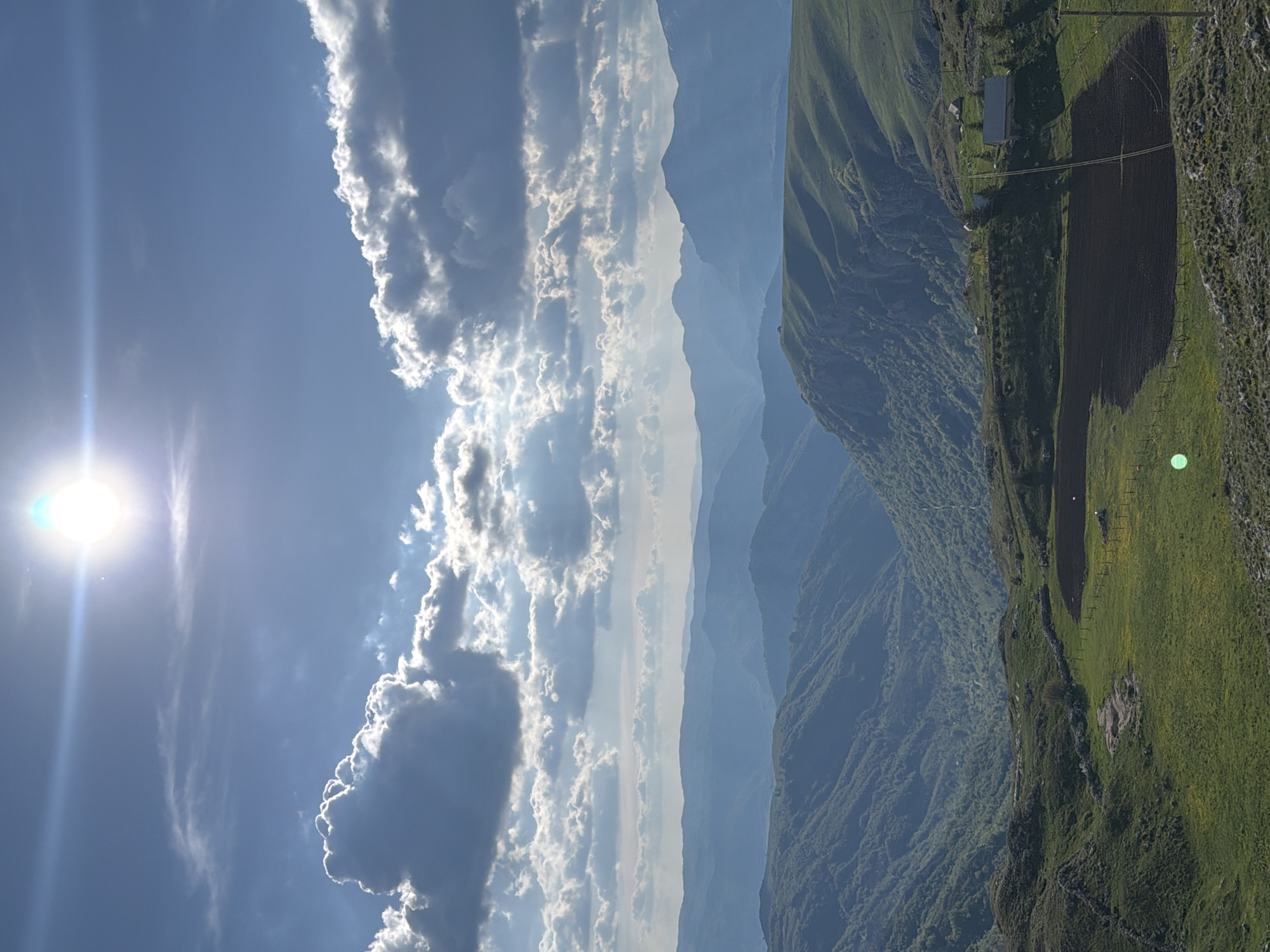 The image shows a landscape near Boričje, Montenegro. In the foreground, there are steep, green, mountainous hillsides with evidence of cultivated fields and a small building. In the mid-ground, there are layers of blue-grey mountains fading into the distance. The sky is a bright blue with puffy white clouds extending vertically, and the sun is visible with a lens flare effect.