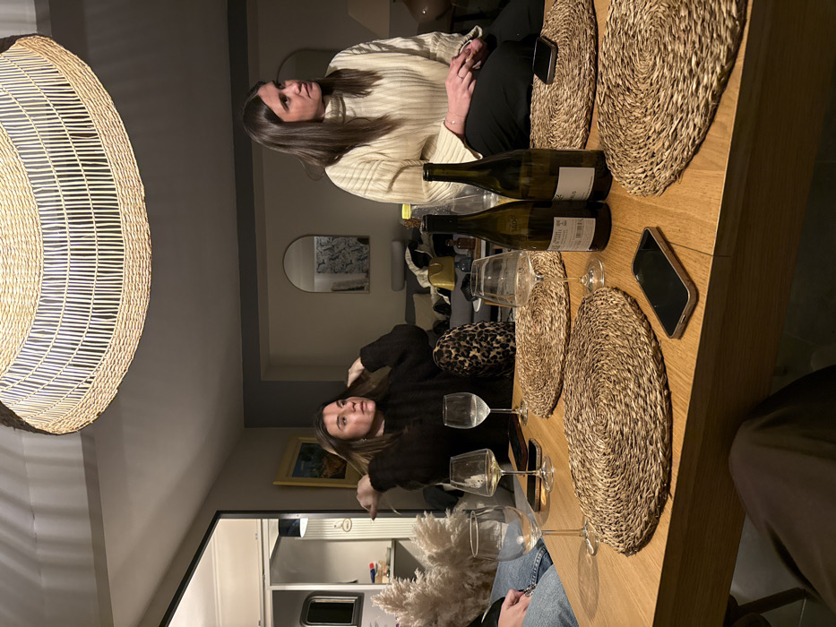 The image depicts an indoor social gathering, likely an evening dinner or drinks, in Saint-Étienne, France. Two women are seated around a wooden dining table in what appears to be a home or restaurant setting.
One woman, with long dark hair, wears a light-colored, ribbed turtleneck sweater and dark trousers. She is seen from the chest up, with her hands clasped in front of her, looking attentively towards the side. The second woman, also with long dark hair, wears a dark, fuzzy top. She is positioned closer to the viewer, looking towards the camera with a neutral expression, with her hands near her head, possibly adjusting her hair.
The wooden table is set with several items. Two empty wine bottles, one green and one lighter, stand prominently. At least four wine glasses are visible, some empty, and one possibly containing white wine. Four round placemats, made of woven natural fibers, are arranged on the table. Two smartphones are placed face down. A decorative element resembling fluffy pampas grass is also on the table.
In the background, a large, circular pendant light fixture made of woven material hangs from the ceiling, providing ambient lighting that suggests it is nighttime. An arched mirror on the wall reflects a patterned background, and a framed piece of art with a yellow border is visible further back. A leopard print cushion or bag rests on a seat next to one of the women.
The scene conveys a relaxed and convivial atmosphere. The text on the wine bottle labels is largely unreadable, though parts of "MOS" and "BLANC" are faintly visible on one.