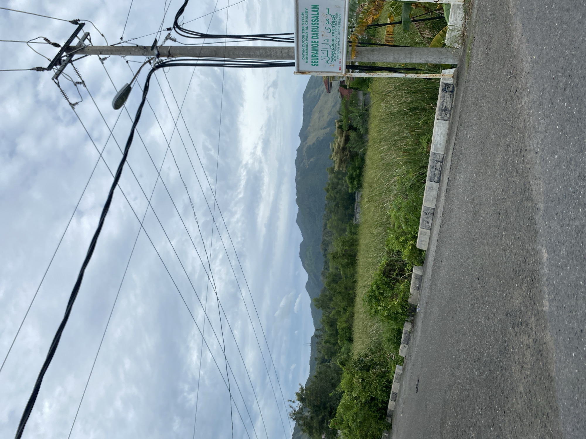 The image shows a rural scene in Beuradeun, Indonesia. A utility pole with power lines dominates the left side of the image. A sign, "SEURAMOE DARUSSALAM," with Arabic script, is mounted on a concrete post to the right of the pole. Vegetation is present along the roadside. In the background, a mountain range is visible under a cloudy sky. A paved road extends into the foreground.