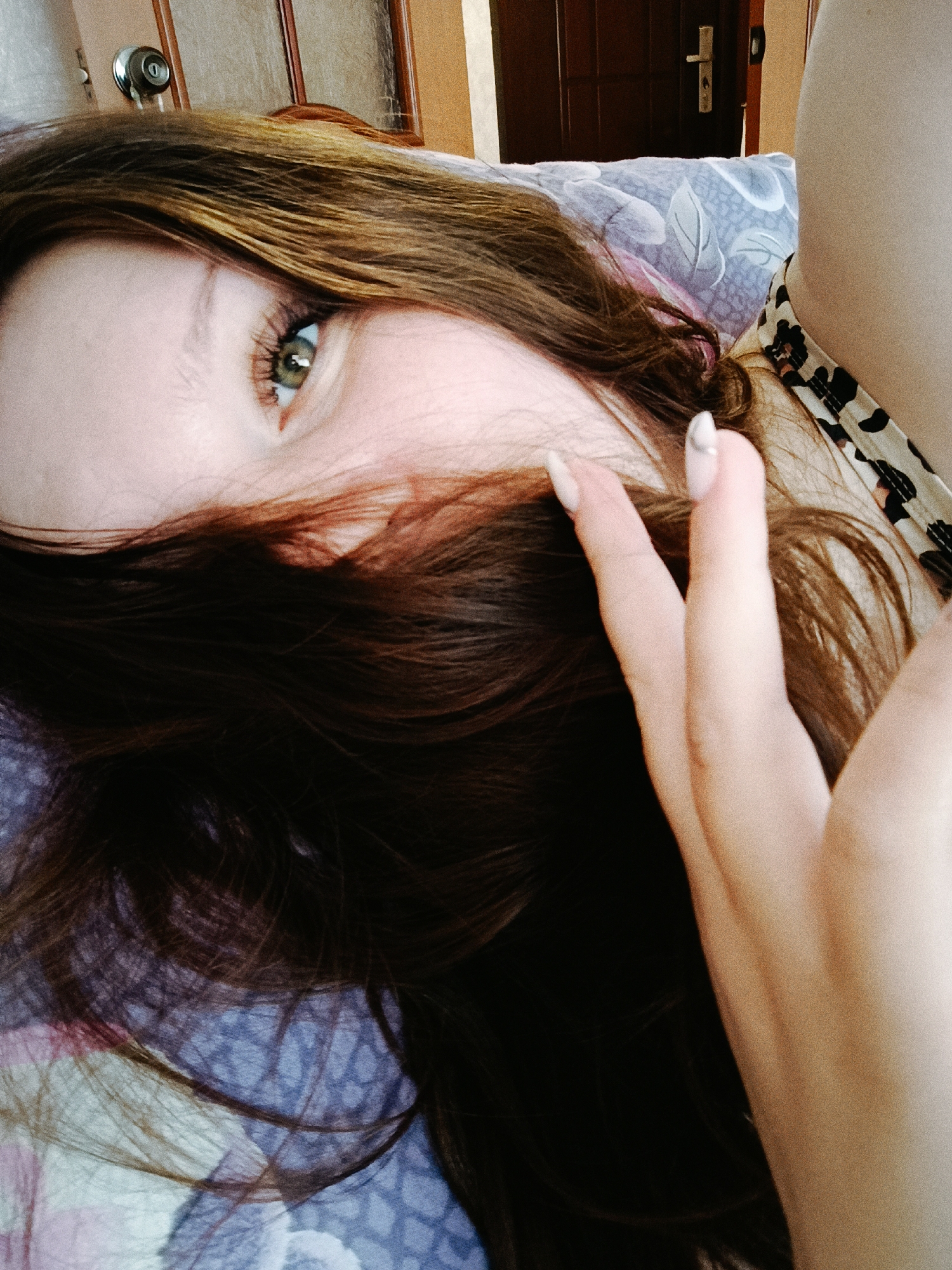 A woman lies on a bed with a blue and purple patterned bedspread, partially covering her face with her brown hair. Her left eye is visible, looking towards the camera. Her right hand is raised, fingers spread, with long nails painted white. She appears to be wearing a garment with a leopard print trim. In the background is a wooden door and a wall. The location is Tobolsk, Russia.