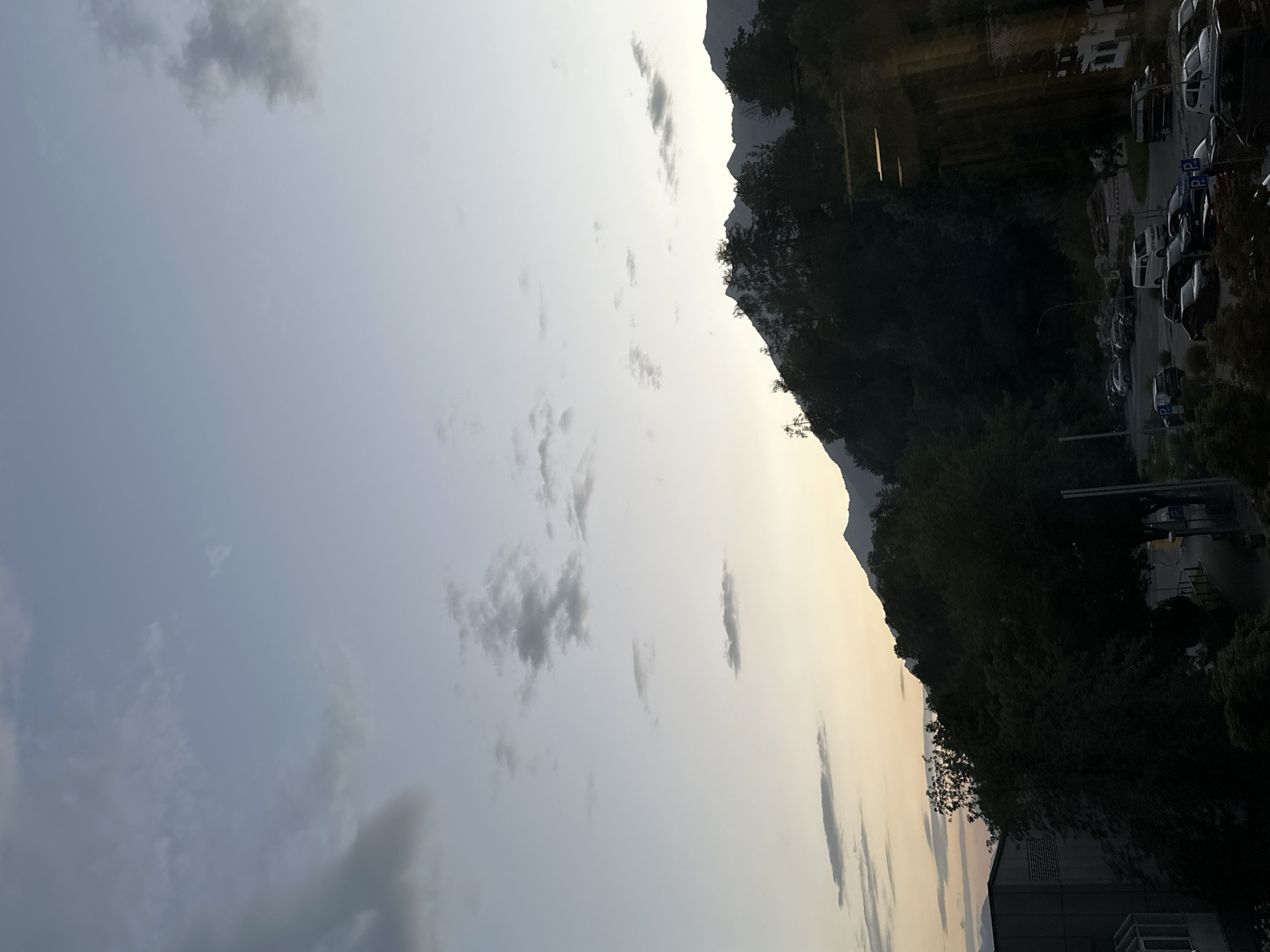 The image shows a view of the town of Garmisch-Partenkirchen, Germany, at dusk. The sky is a pale blue with scattered clouds, and the sun is setting behind a line of silhouetted mountains. In the foreground, there is a dense forest of dark green trees. Below the forest, there is a parking lot filled with various cars, including sedans, SUVs, and vans. A few streetlights are visible, illuminating parts of the parking lot and an adjacent road. To the left, a modern building with a grey facade and visible air vents can be seen. The overall impression is one of a peaceful, mountainous town at the end of the day.