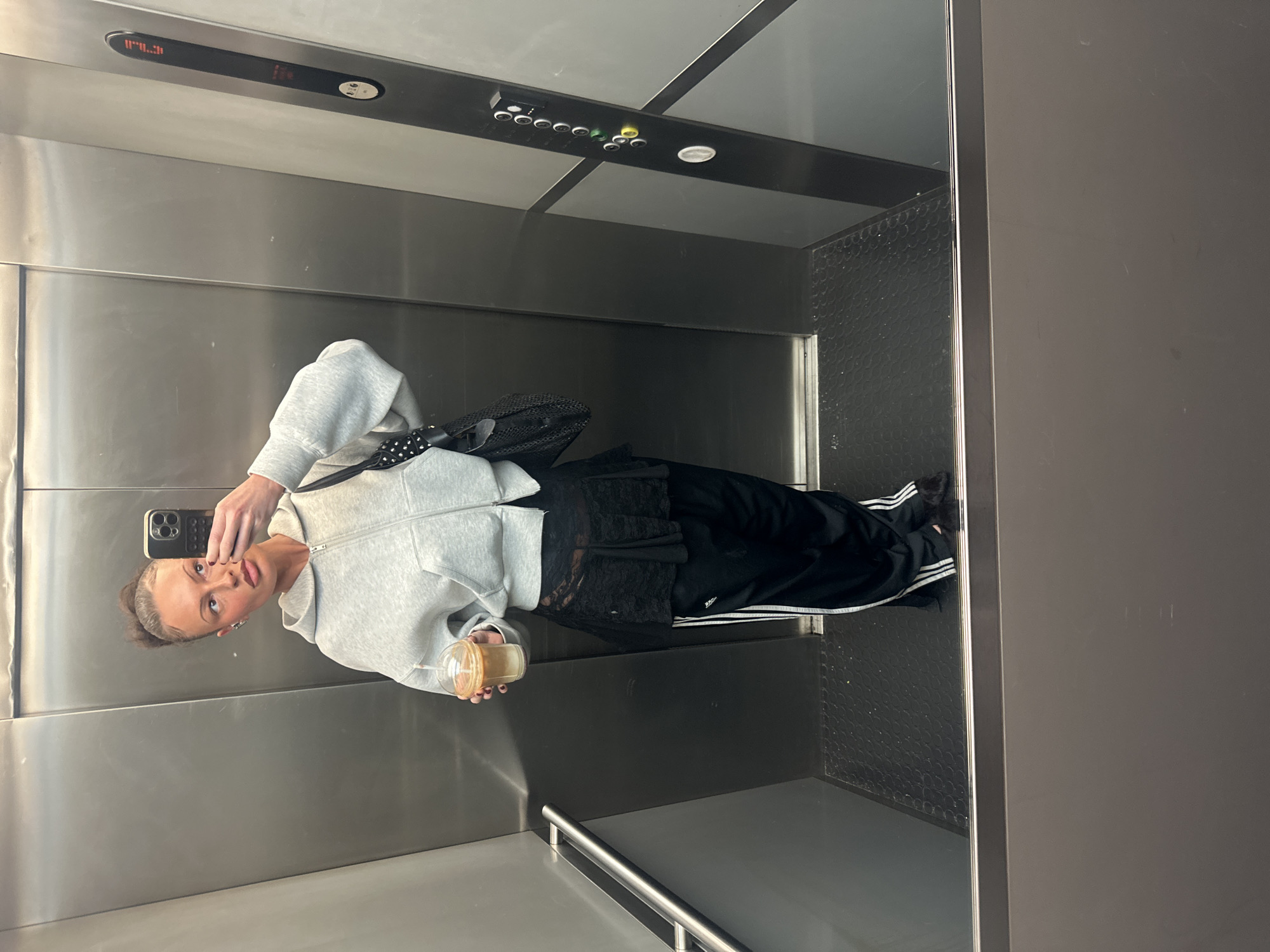 A woman is taking a selfie in an elevator with a mirrored interior. She is wearing a grey zip-up hoodie, black pants with white stripes, and holding a clear plastic cup with a brown liquid inside. She is facing the mirror, holding a phone with multiple lenses visible on the back. The elevator has a panel of buttons and a digital display above. The elevator floor appears to have a textured surface. The environment is reflective due to the mirrored walls. No information suggests this is located in London, United Kingdom.