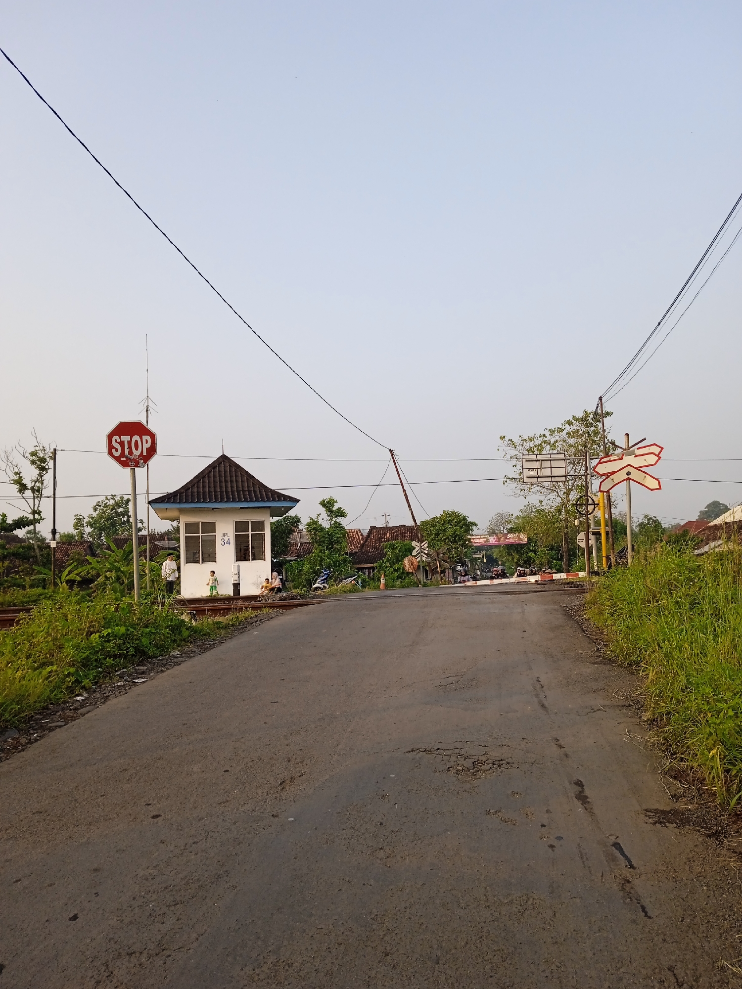 The image depicts a rural road crossing a railroad track in Sidorejo, Indonesia. A small, white control booth with a black roof is located to the left of the road, adjacent to a stop sign. A railroad crossing sign stands to the right of the road. Vegetation lines both sides of the road. People are present near the control booth and on the railroad track. Several motorcycles are present beyond the gate. The road itself is asphalt, showing signs of wear and tear. Overhead power lines extend across the scene.