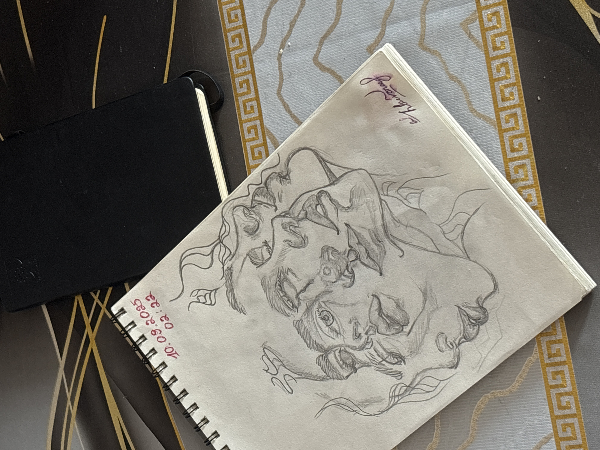 This still life image, captured in Yerevan, Armenia, features a white spiral-bound sketchbook open to a pencil drawing, positioned on a patterned surface. Alongside it rests a closed black notebook or journal. The underlying surface displays two distinct patterns: one section is black with thin, abstract golden lines, while the adjacent area is light gray/beige, adorned with a golden Greek key (meander) border and subtle wavy golden lines.
The central subject is the intricate pencil sketch within the open sketchbook. It depicts two stylized human faces, seemingly intertwined or in close proximity, surrounded by flowing lines that suggest hair or dynamic energy. The faces are rendered with expressive details, including visible eyes and lips, conveying a sense of intensity or deep connection. The artistic style is distinct and evocative.
Handwritten details are visible on the sketchbook page. In the bottom left corner, in red ink, the date "10.09.2085" and time "02:32" are inscribed, indicating a future date and an early morning hour. In the top right corner of the drawing, a signature or artistic tag in purple ink, possibly reading "K. Kevoronef," adds a personal touch. The black notebook is plain and closed, contrasting with the detailed artwork. The overall scene suggests an indoor setting, possibly a desk or table where creative work takes place.