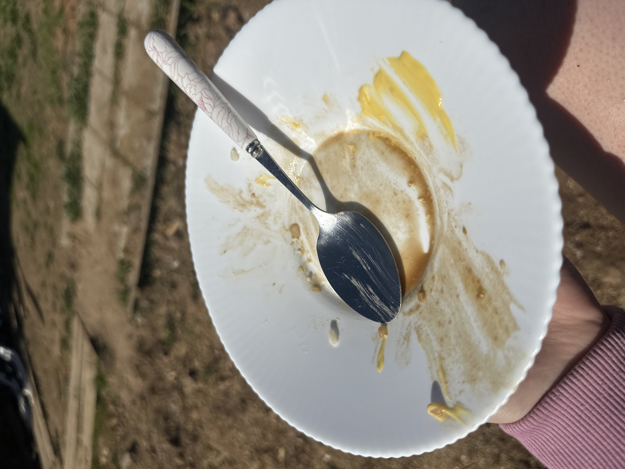 The image depicts a white, scalloped-edge plate with remnants of food, including a smear of yellow and a ring of brown, presumably from a finished meal. A silver spoon with a white floral-patterned handle rests on the plate. The plate is held by a person, as indicated by the presence of a forearm clad in a pink, ribbed sweater. The background is blurred, suggesting an outdoor setting with brown and green hues. There is no information about the city of Черёмушки, Russia, in the image.
