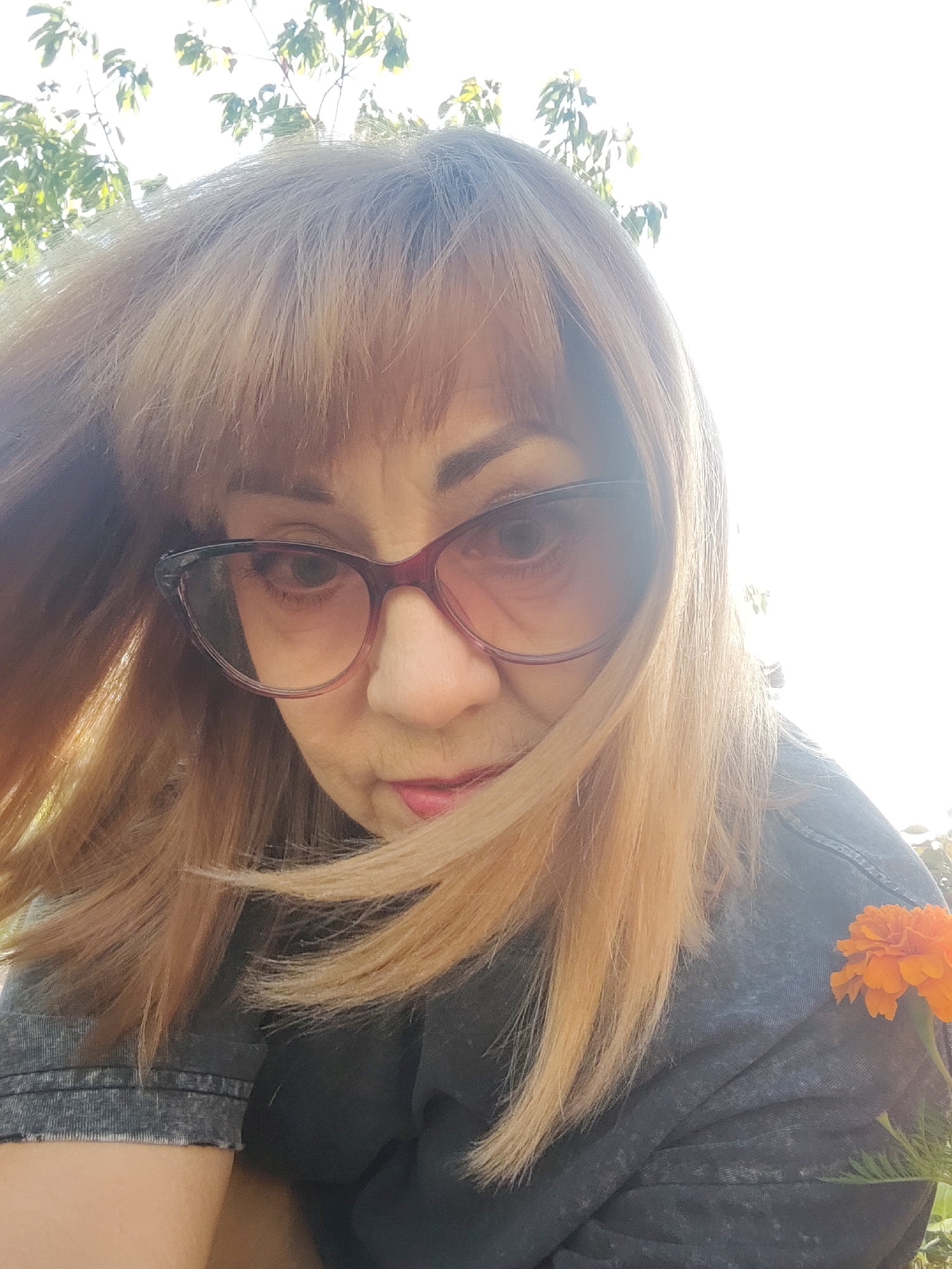 A woman with shoulder-length blonde hair and bangs is looking down and to the left, framed by her hair. She is wearing large, reddish-purple cat-eye glasses, and a dark grey t-shirt with a faded, mottled pattern. An orange marigold is pinned to her t-shirt on the right side of the frame. The background is blurred with bright sunlight, suggesting an outdoor setting with trees overhead. There is no discernible urban environment.