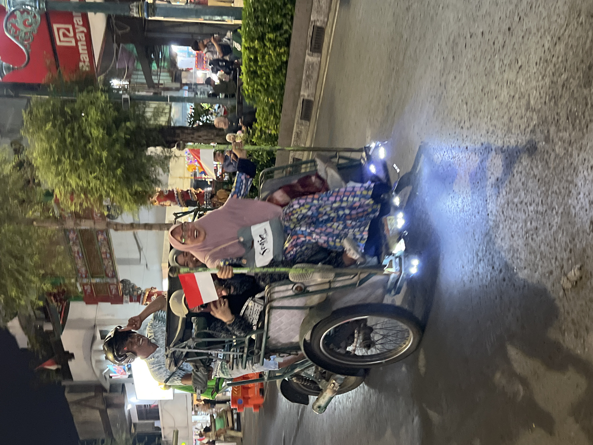 In this nighttime image captured in the city of Yogyakarta, Indonesia, a three-wheeled passenger vehicle, commonly known as a becak, navigates a street. The becak is occupied by at least four individuals. The driver, wearing a helmet, is at the front, appearing to gesture with his left hand. Behind him, a person holds a small Indonesian flag, red and white. Two passengers are seated behind the flag holder. The passenger on the right, a woman wearing a pink headscarf, is smiling broadly and looking forward. She is carrying a light blue bag with the word "Sojuu" written on it in black script. The becak is equipped with bright, possibly blue, lights illuminating the road beneath it. In the background, illuminated storefronts and foliage are visible, suggesting a commercial or bustling urban area. The street surface is asphalt, with some reflections indicating it might be wet.