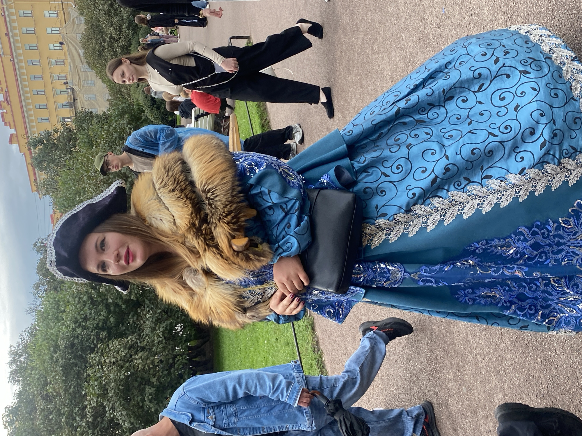 A woman in a blue dress with a fur collar and a pirate hat walks down a path in a park in Saint Petersburg, Russia. The dress is embellished with intricate blue and silver patterns, and the fur collar is made of brown and tan fur. The woman is carrying a black handbag. In the background, other people are walking and sitting on benches. There are trees and buildings visible.