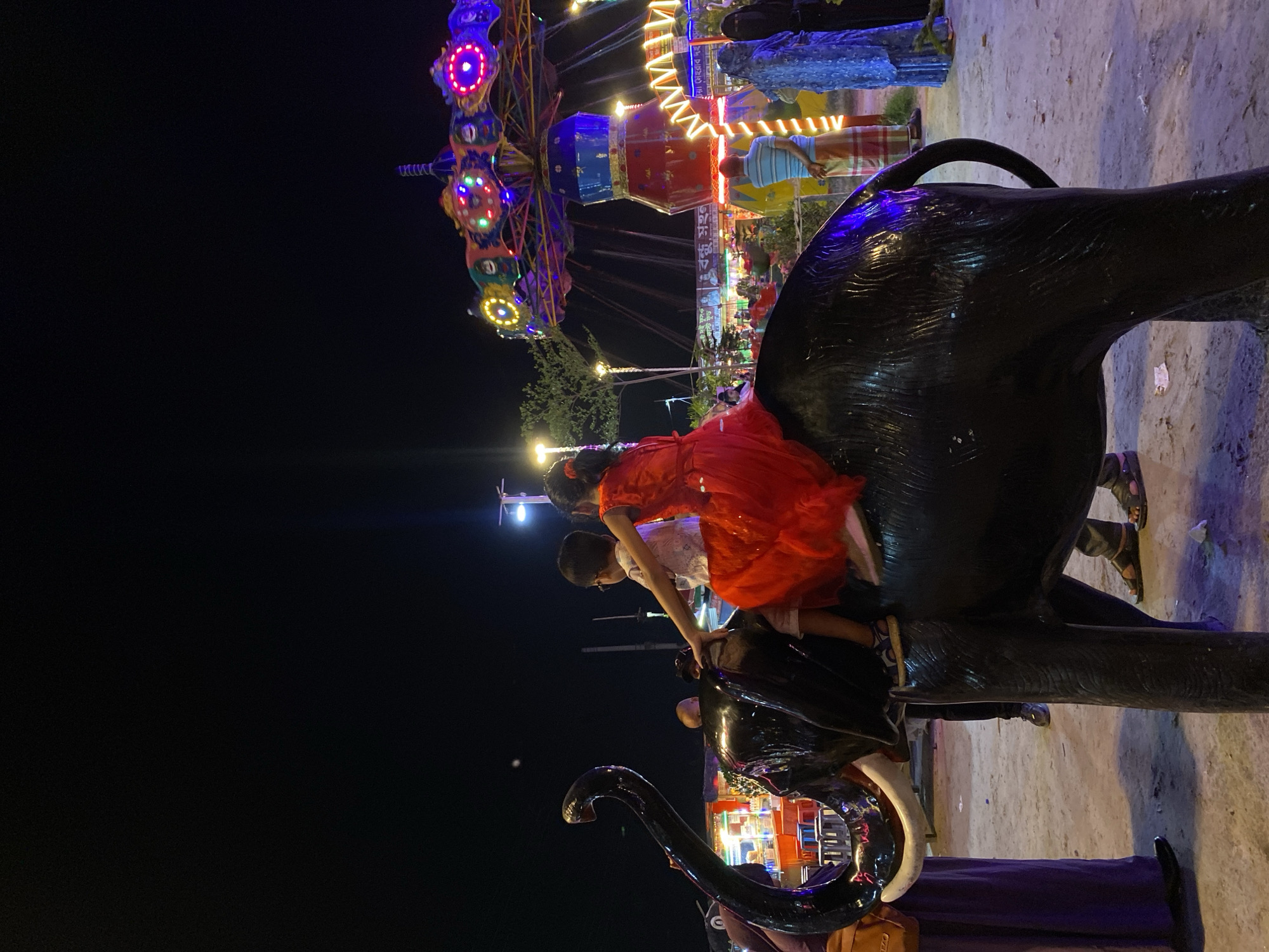 At night in Dhaka, Bangladesh, a fairground scene unfolds with illuminated rides visible in the background. A group of people, including two children, are interacting with a large, dark-colored, glossy statue of an elephant. One child, wearing a bright red dress, is perched on the elephant's back, while another child in a light-colored shirt reaches towards the elephant's head. The environment is lit by fairground lights and street lamps. Other individuals are standing around the base of the statue.