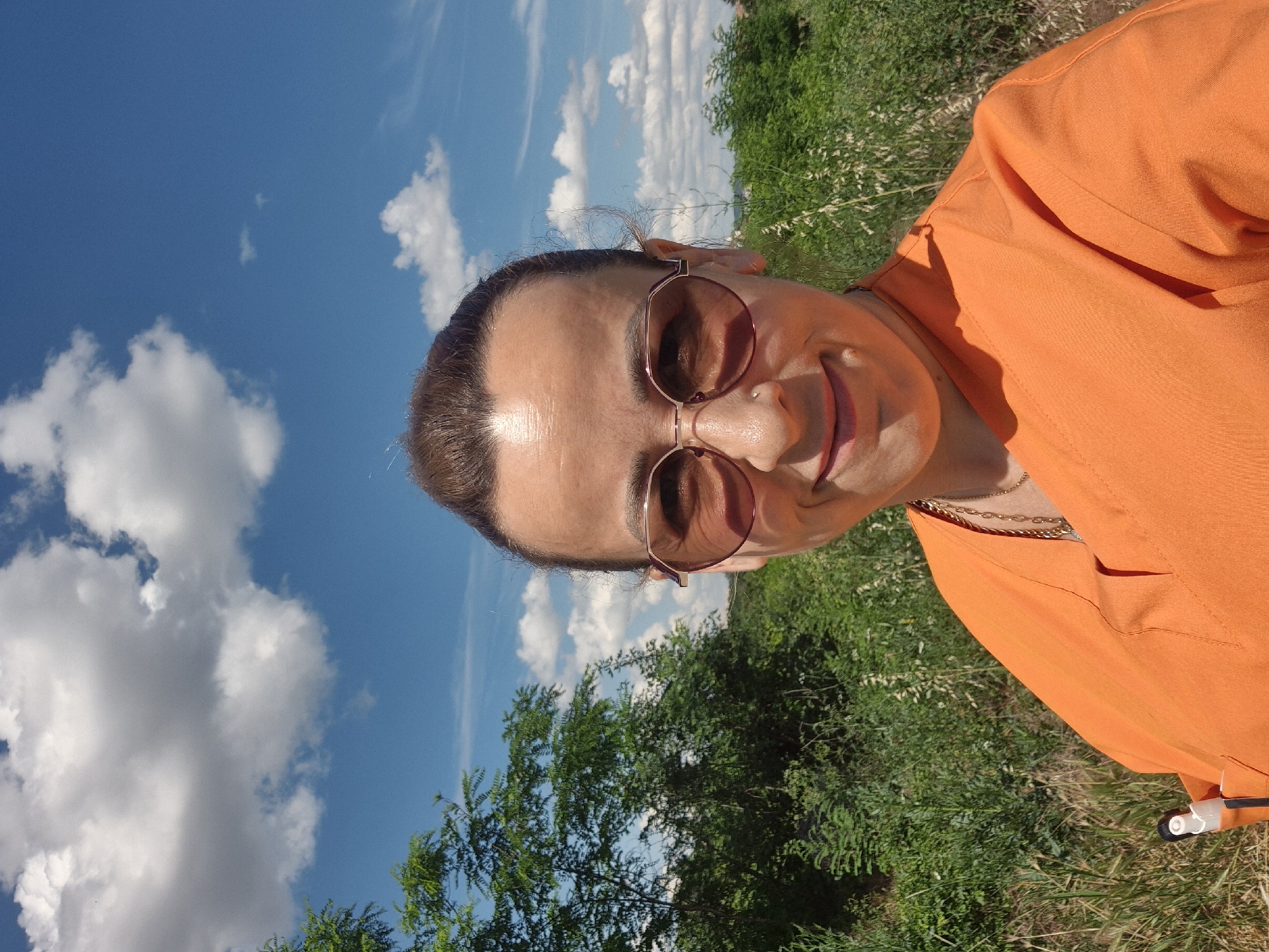 A woman with brown hair pulled back and wearing rose-tinted aviator sunglasses and an orange scrub top is taking a selfie outdoors on a sunny day with blue skies and white clouds. She is smiling and looking at the camera. Her left shoulder and chest are visible, as well as a portion of the surrounding vegetation. A white object, possibly a pen, is tucked into the chest pocket of her scrub top. The background consists of green trees and bushes under a bright blue sky dotted with fluffy white clouds. The location is described as Unknown, Unknown.