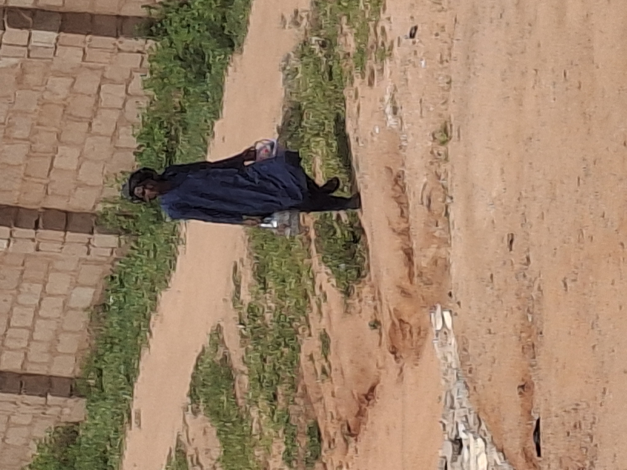 A person walks on a dirt path next to a brick wall. The person is wearing a dark blue robe and headscarf and is carrying a red and white container in their right hand and a clear plastic bottle in their left hand. The path is lined with sparse green vegetation. The image appears to have been taken in the city of Tumfure, Nigeria.