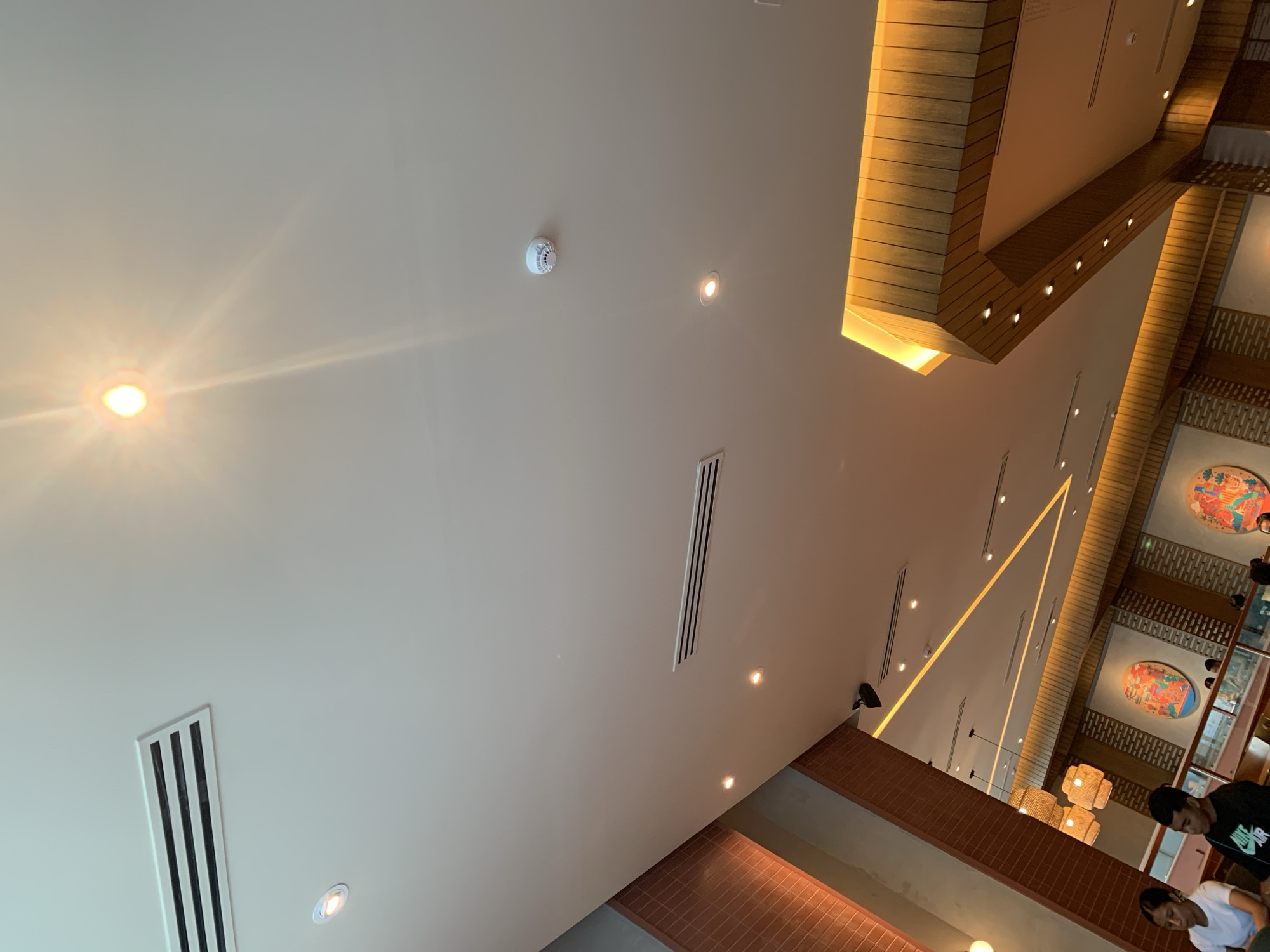 The image depicts the interior of a restaurant in Angeles City, Philippines. The perspective is from a low angle, looking up at the ceiling. The ceiling is white and features several recessed lights and air conditioning vents. A white circular smoke detector is also visible on the ceiling.
The restaurant has a modern design with warm lighting. On the right side of the image, a section of the ceiling is illuminated with a golden hue, highlighting a wooden slatted structure. In the background, decorative round wall art with colorful depictions is visible.
Two young people, a female and a male, are seated at a table in the foreground on the left. The female is wearing a white t-shirt, and the male is wearing a black t-shirt with a white logo. Other patrons are visible in the background, suggesting a busy dining environment. The overall ambiance appears to be casual and inviting.