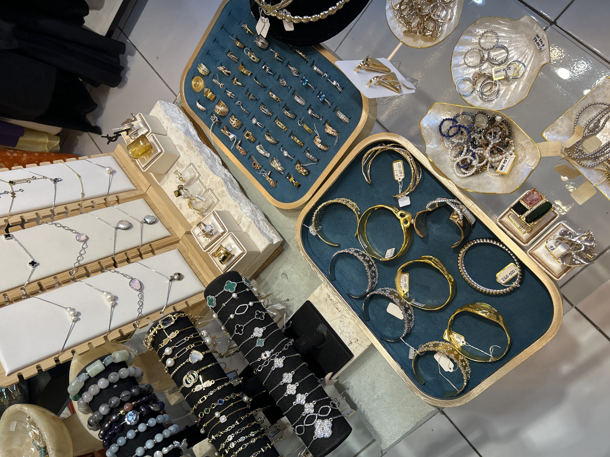 An overhead shot showcases a collection of jewelry displayed on glass surfaces and pedestals within a retail environment. The jewelry includes necklaces, bracelets, and rings, featuring a variety of materials such as silver, gold, pearls, and gemstones. The display is organized with multiple trays and stands, some of which are lined with teal velvet. Several price tags are visible, indicating the items are for sale. The overall impression is one of an organized and well-stocked jewelry store, likely located in a commercial area. Without further information, the specific location within Ubud District, Indonesia cannot be definitively determined from the image alone.