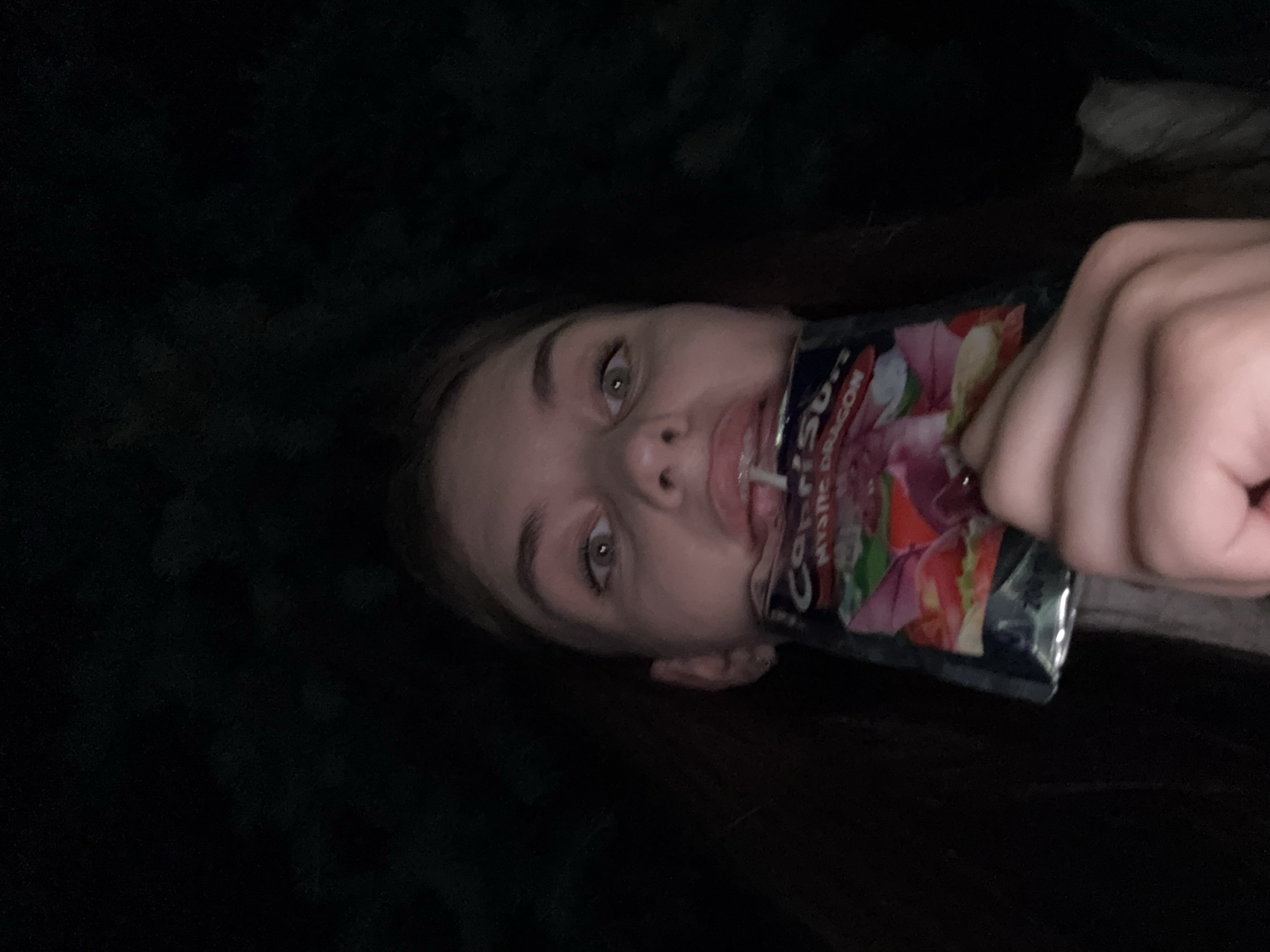 A young person, possibly female, is shown from the chest up, holding a pouch of Capri Sun "Mystic Dragon" juice near their mouth in an outdoor, dark environment. The person's face is partially obscured by the pouch, but their eyes are wide open and looking directly at the camera. There is some liquid on their lips. The background is dark and indistinct, with hints of trees or foliage. The image appears to be a selfie taken at night. There is no indication of the location being the city of Dnipro, Ukraine.