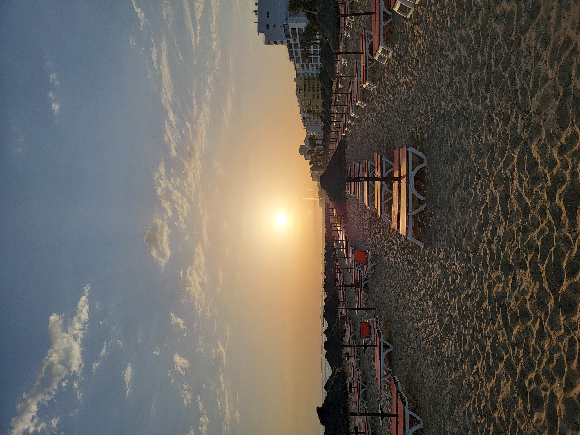 The image depicts a serene beach scene in Quarteira, Portugal, during what appears to be sunrise or sunset. Rows of unoccupied lounge chairs with thatched umbrellas are neatly arranged on the sandy beach, stretching towards the horizon. The sun, a bright orb, is visible in the sky, casting a warm glow over the scene. In the background, a line of white and colored buildings, typical of a coastal resort town, forms the skyline. The sky is a soft blue, dotted with scattered white clouds. The sand exhibits textured patterns, indicating the tide or wind. The overall atmosphere is peaceful and still, with no people or animals present.