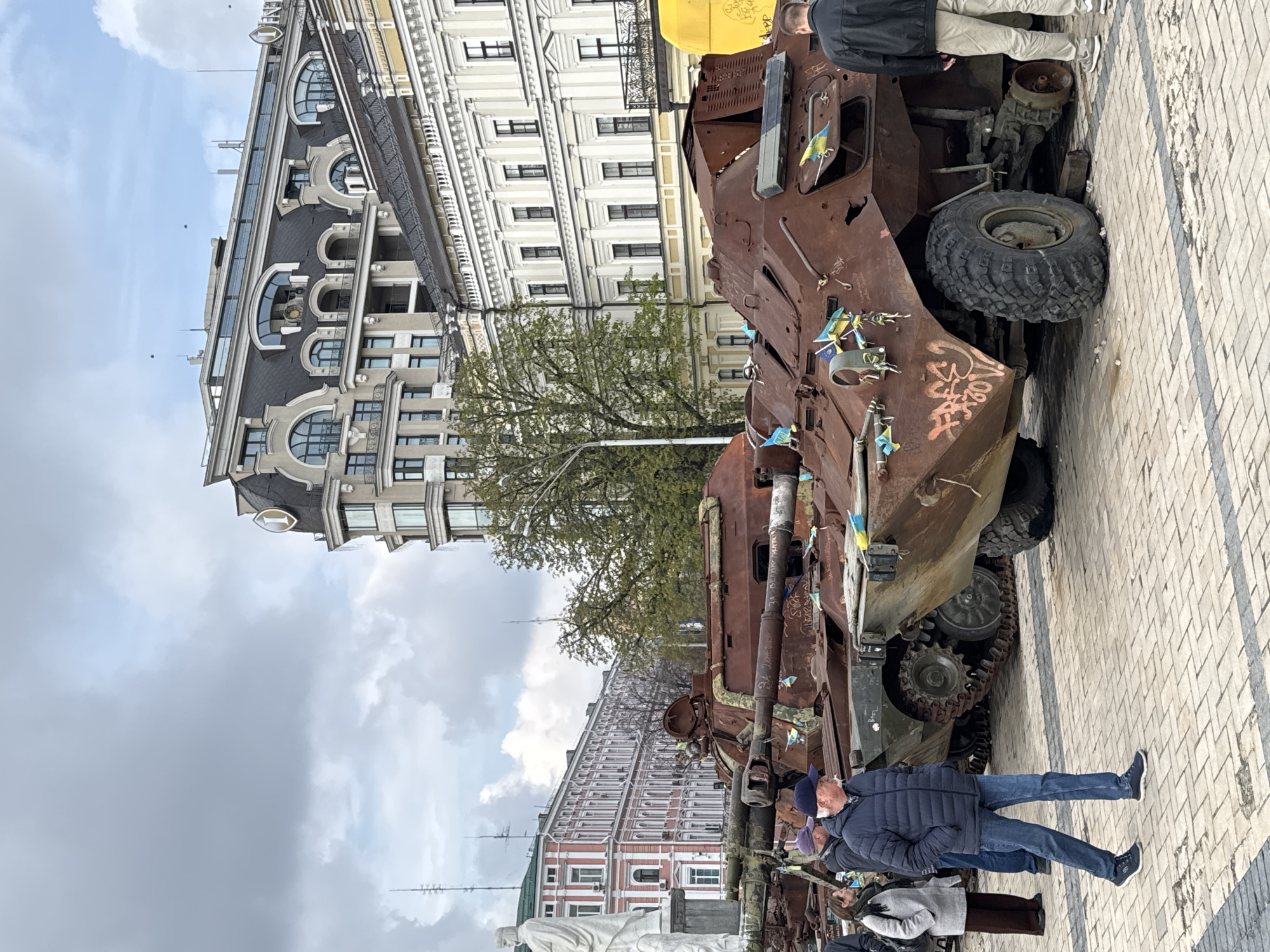 The image shows a destroyed Russian military vehicle on display in Kyiv, Ukraine. The vehicle, likely an armored personnel carrier, is heavily damaged, with signs of rust and graffiti. Small Ukrainian flags are attached to it. People are standing nearby, observing and interacting with the vehicle. In the background are buildings characteristic of Kyiv's architecture. The sky is overcast. The ground is paved with a pattern of light and dark gray bricks.