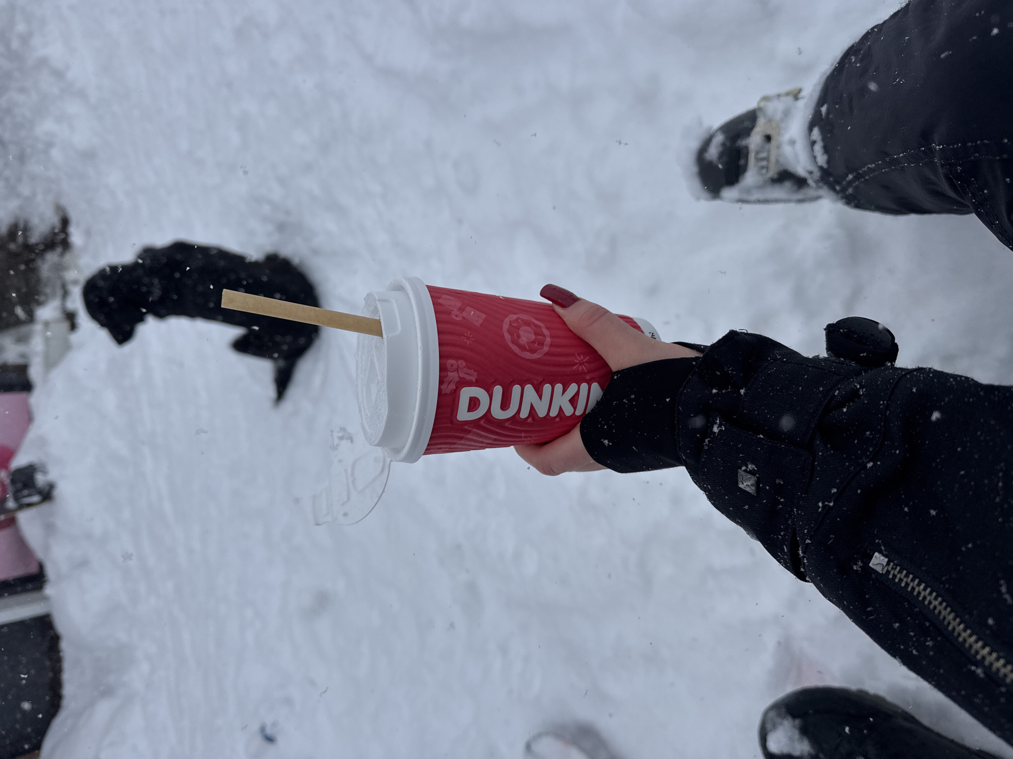The image shows a person standing in snow, holding a Dunkin' coffee cup. The environment is covered in snow. The person is wearing black winter clothing and black boots. The Dunkin' cup is red and has a white lid with a stirrer. The nails of the hand holding the cup are painted red.