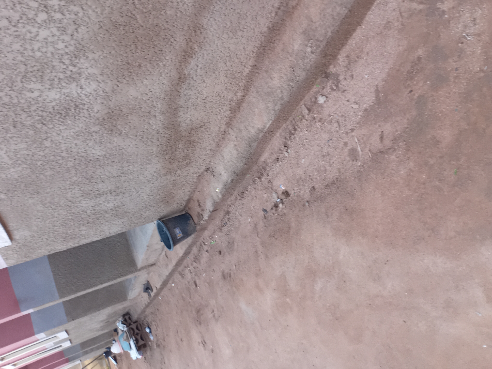 The image depicts an outdoor scene, likely in Jauro Yinu, Nigeria, with a focus on the ground and the side of a building. A dark-colored bucket is visible near the wall, positioned on a patch of dirt and debris. The building's wall has a textured, rough finish. The ground is a mix of dirt, sand, and small stones, with a shallow trench or rut running parallel to the building. In the background, a section of the building's exterior shows a pink and gray color scheme, with what appear to be window openings. A few individuals are partially visible in the far left background, near some scattered objects on a bench-like structure. The overall impression is of an unpaved, utilitarian outdoor space.