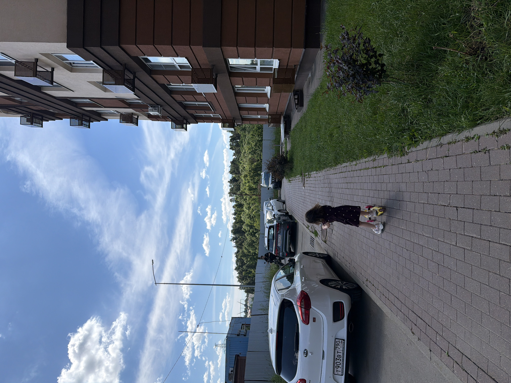 In Lopatino, Russia, on a clear day with a bright blue sky and scattered white clouds, a young girl in a dark patterned dress rides a yellow scooter down a brick-paved sidewalk. To her left, a white Kia hatchback is parked along the street, facing away from the camera. Behind the Kia, several other cars are parked in a row, including a dark grey SUV. Adjacent to the sidewalk is a well-maintained grassy area. On the right side of the image stands a modern, multi-story residential building with a brown and cream facade. The building features multiple windows and air conditioning units mounted on the exterior. A black metal fence encloses the area behind the parked cars and along the edge of the property. In the background, a dense line of green trees forms a natural barrier. A street lamp with a curved arm is visible in the distance.