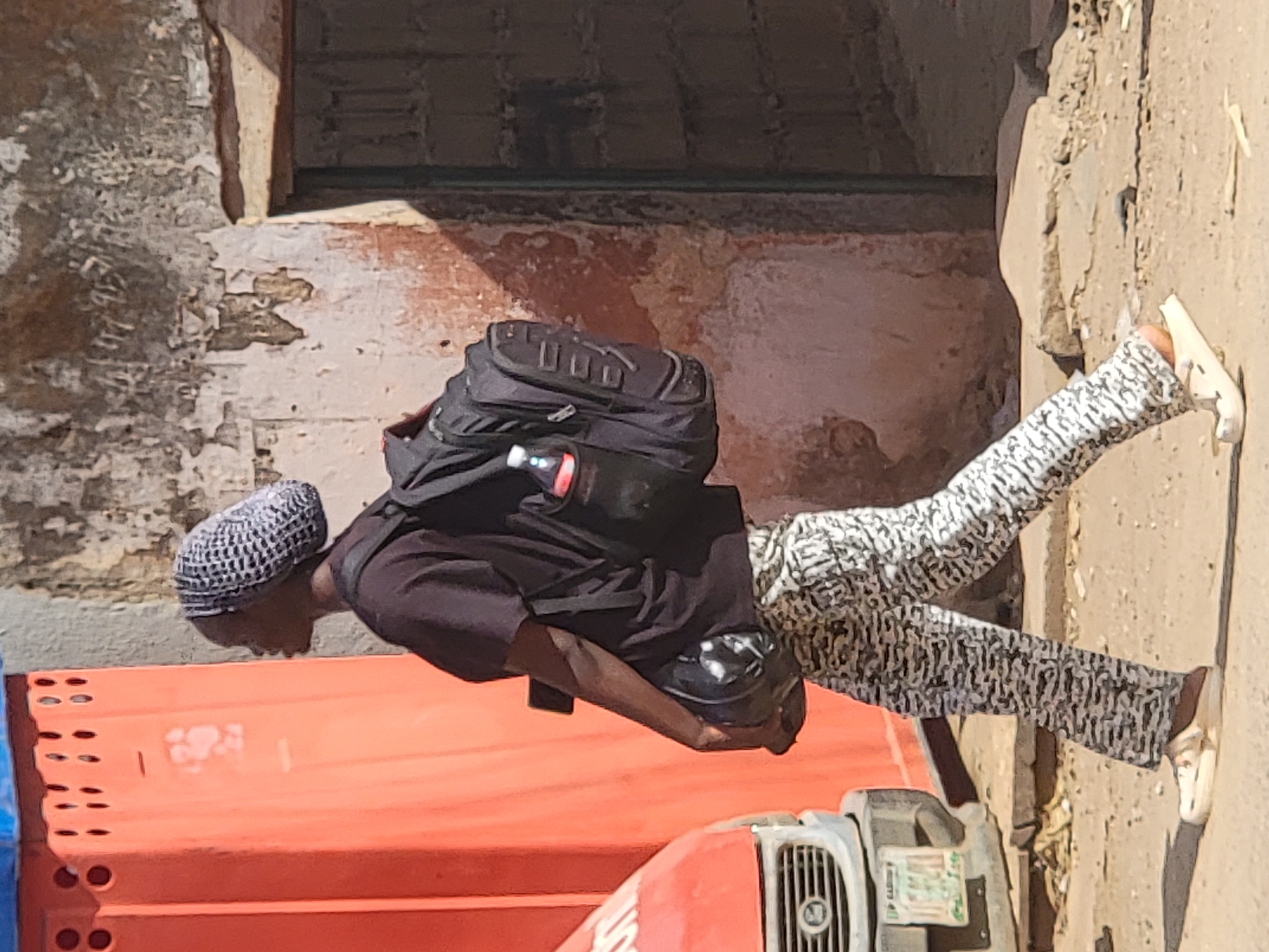 A man with dark skin and a patterned cap is standing next to a vehicle on a sunlit street in Maiburiji, Nigeria. He is wearing a black shirt, patterned pants, and white sandals. He has a black backpack with a water bottle attached to it. He appears to be standing near a building with weathered walls and a door. A portion of an orange and red vehicle is visible in the lower part of the frame. The ground is dusty and uneven.