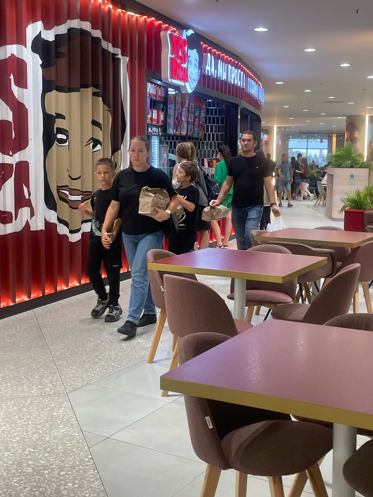 In a food court in Sochi, Russia, a woman in a black t-shirt and blue jeans walks with two young boys, one on her left and one on her right. She is holding a brown paper bag in her right hand. The boy on her left is wearing a black t-shirt and black pants. The boy on her right is wearing a black t-shirt. Behind them, a man in a black t-shirt and blue jeans walks away from the camera. In the background, there is a food establishment with a large mural of a cartoon face on its exterior wall. The establishment has red vertical panels and a sign with "YES PIZZA" written on it. In the foreground, there are several tables and chairs, some with pink tops and brown fabric seats. Other people are visible in the background, walking through the food court. The floor is tiled with a light gray pattern.