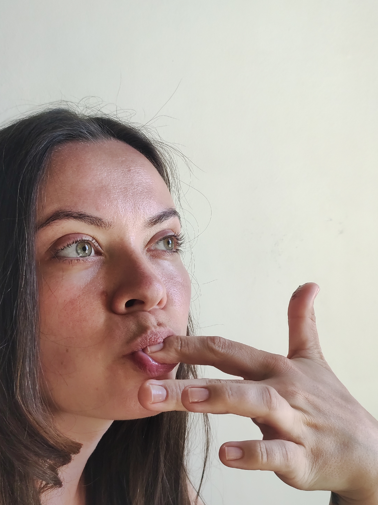 A woman with long brown hair and green eyes is looking to the upper left. She has her fingers in her mouth and is possibly licking them. The background is a plain off-white wall.