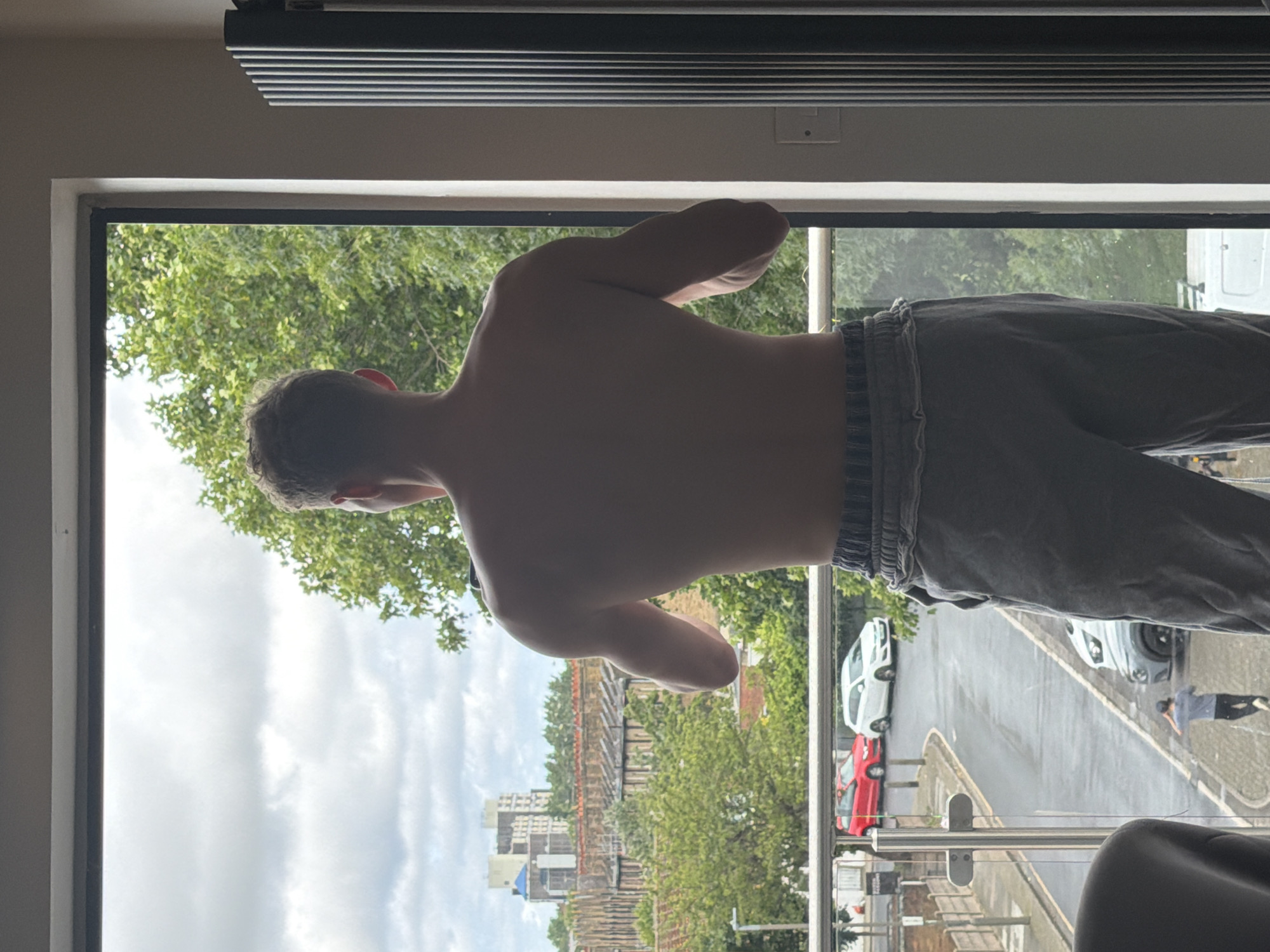 A man stands with his back to the camera, looking out a large window. He is shirtless and wearing grey sweatpants. The window overlooks a street with parked cars and some buildings in the distance. Trees are visible outside the window. The scene appears to be in an urban setting, possibly London, United Kingdom, given the architectural style of the buildings and the presence of a red car and a white car, common car models in the UK. The man's posture suggests he is contemplating the view. There are no explicit actions or interactions depicted, but the overall impression is one of quiet observation.