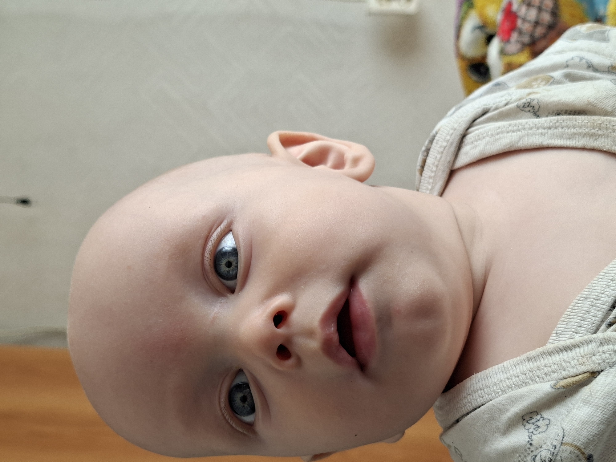 A close-up, waist-up shot captures a bald baby looking slightly to the right of the camera. The baby has large, blue eyes, a small nose with red nostrils, and an open mouth with visible lips and a hint of the tongue. A slight redness is present on the baby's forehead, and one ear is visible on the right side of the face. The baby is wearing a light-colored onesie with a subtle pattern of outlined drawings. The background consists of a soft, muted texture with an orange curtain on the left and a wall with a faint pattern on the right. A blurred, colorful object, possibly a stuffed toy, is partially visible on the right side of the frame. The lighting is soft, casting subtle shadows. The scene appears to be indoors, in an unknown location within an unknown city.