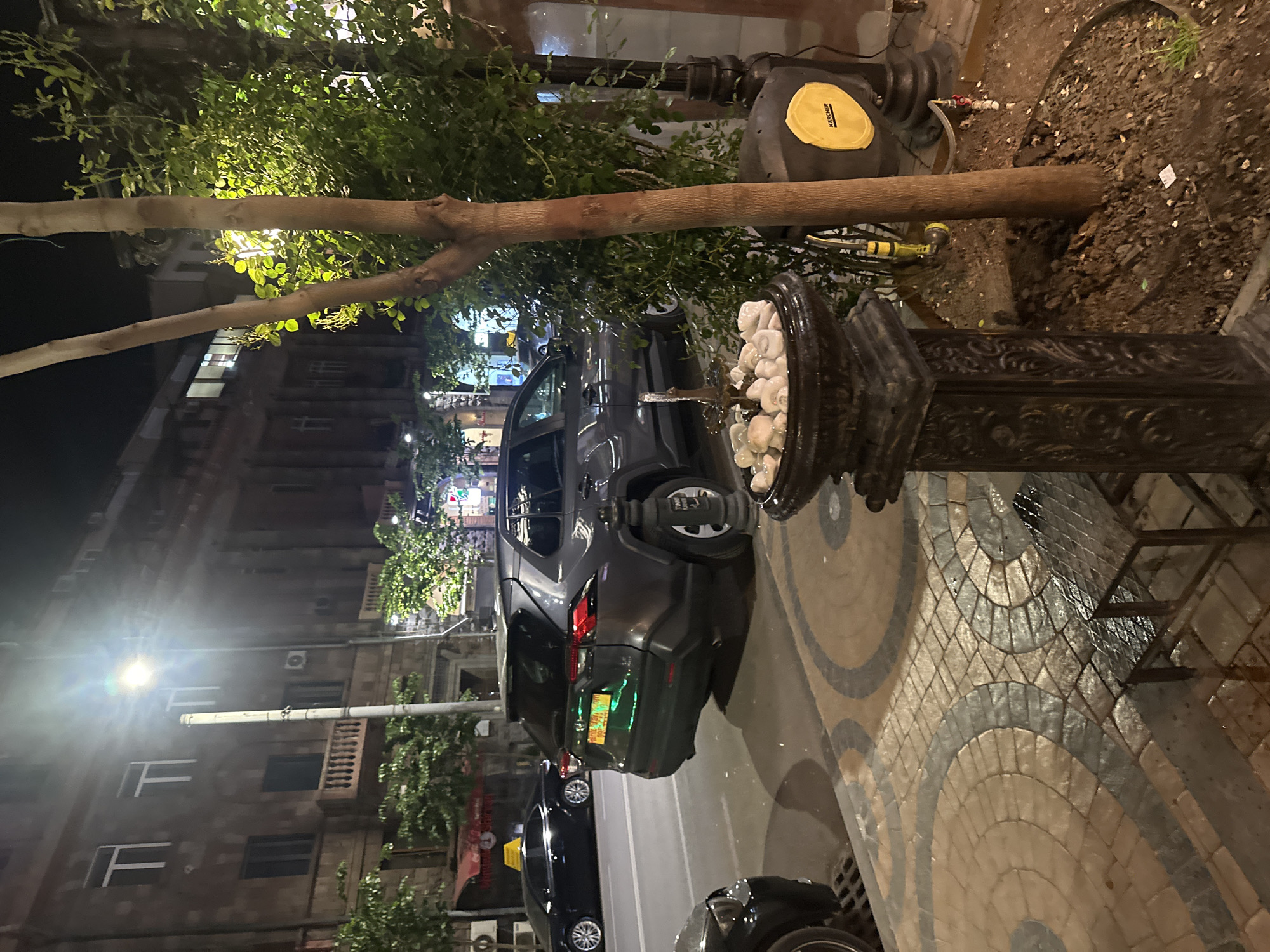 This image, likely taken at night, shows a street scene in Yerevan, Armenia. In the foreground is a decorative water fountain. Behind the fountain, a gray SUV with a partially visible license plate that may read "800 TA 200" is parked on the street. Other cars are parked further down the street. The road is paved with a patterned design. A tree branch extends across the top portion of the image, partially obscuring a black water hose reel with a yellow sticker. Buildings with multiple windows line the left side of the street, and streetlights illuminate the scene. The leaves from the trees partially cover the building, and they also cover the parked car to some extent. The building appears to be made of red brick, and there are multiple layers of windows.