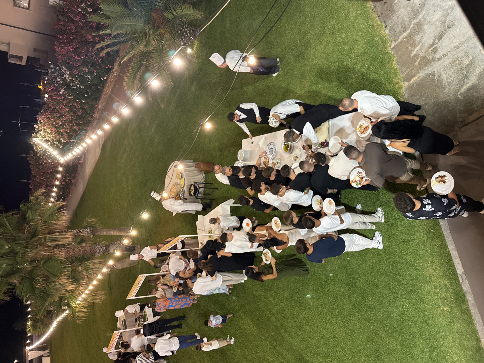 This aerial image depicts an outdoor evening event with a buffet setup on a lush green lawn in Isola delle Femmine, Italy. String lights are draped across the scene, illuminating the gathering. A group of people, dressed in various attire including chefs' uniforms and formal wear, are congregated around tables, being served food. Several individuals are holding plates with food, suggesting they are either serving themselves or have just received their meals. The environment features palm trees and a building in the background, consistent with a coastal location in Sicily. The scene appears to be a catered event, possibly a wedding reception or a party, taking place on a well-maintained lawn.
