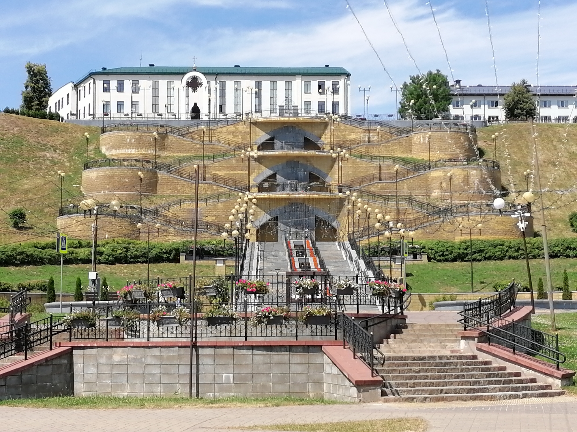 This media file depicts an outdoor scene on a sunny day, likely in the late morning or early afternoon given the bright sunlight and shadows. The setting appears to be a public park or a civic area, characterized by a large, ornate stone staircase and fountain structure leading up to a white building.
The focal point of the image is the elaborate tiered staircase, constructed with stone steps and flanked by curved, terraced walls. These terraces are adorned with numerous lampposts, creating a visually striking effect. At the base of the stairs, a decorative black metal fence encloses planters filled with colorful flowers, adding vibrancy to the scene. The staircase itself has a central section with what appears to be a water feature or a decorative element that cascades down.
Behind the staircase, on the elevated ground, stands a long, white building with a green roof. The building has multiple windows and an arched entrance with decorative metalwork, topped with a cross, suggesting it might be a historical or religious institution. To the left and right of this central structure, other buildings with similar architectural styles are visible, suggesting a larger complex.
The landscape is lush with green grass and manicured bushes surrounding the staircase and buildings. The sky is a clear blue with a few scattered white clouds, indicating fair weather. The overall impression is one of a well-maintained and attractive public space, possibly designed for ceremonial or recreational purposes. There is no visible text in the image.