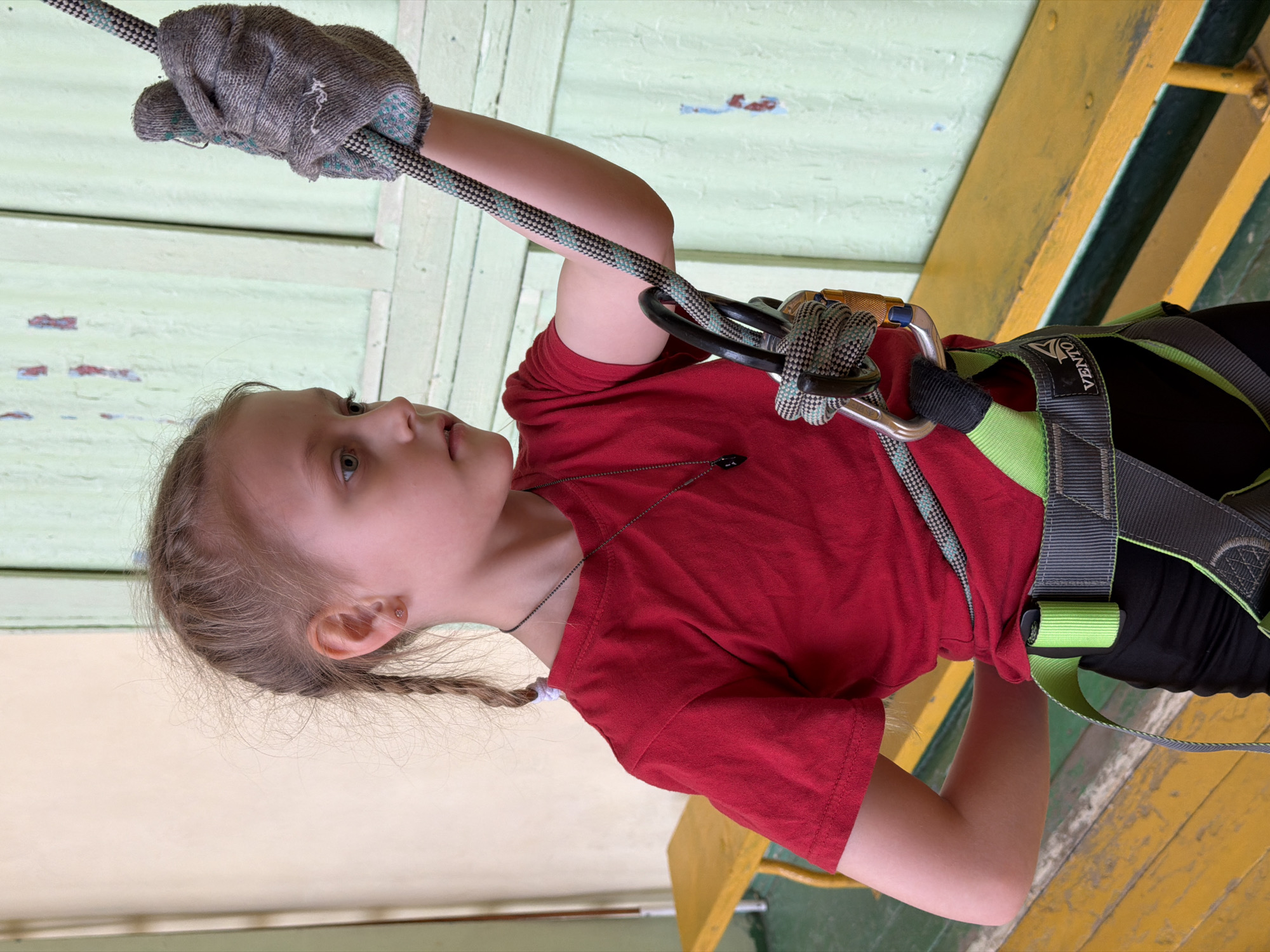 A young girl is depicted mid-activity, likely participating in a rope course or climbing exercise. She has light-colored hair, partly styled in braids, and a focused expression as she looks upwards. She wears a red short-sleeved t-shirt, black pants, and a green and grey full-body safety harness with the brand name "VENTO" visible on a strap. A thin chain necklace and a small stud earring are also visible.
Her hands, covered in grey knitted gloves with speckled palms, grip a thick grey rope dotted with light blue and white. The rope passes through a large metallic carabiner, which is attached to her harness. The carabiner has a black section.
The setting appears to be an indoor or covered outdoor facility, characterized by worn wooden structures. Above the girl, there's a light green wooden wall or ceiling with peeling paint. Below her, bright yellow wooden beams or planks, also showing signs of wear with peeling paint, are visible, alongside some darker green sections. The lighting suggests an ambient, possibly artificial or shaded, environment. The overall impression is one of concentration and physical engagement in an adventure activity.
