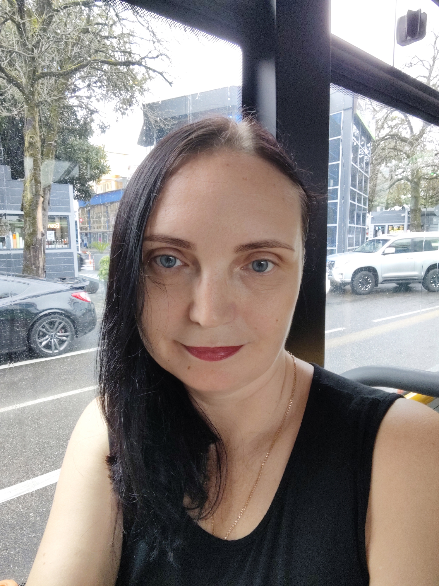 An adult Caucasian woman with dark hair and blue eyes takes a selfie from inside a vehicle. She is wearing a black sleeveless top and a gold chain necklace. Her lips are painted with a dark red lipstick. Through the vehicle's window, a tree with sparse branches and a gray car are visible on a street. In the background, modern buildings and a white SUV are seen. The weather appears overcast, with visible raindrops on the window. The scene is set on a city street in Sochi, Russia, likely during daytime.