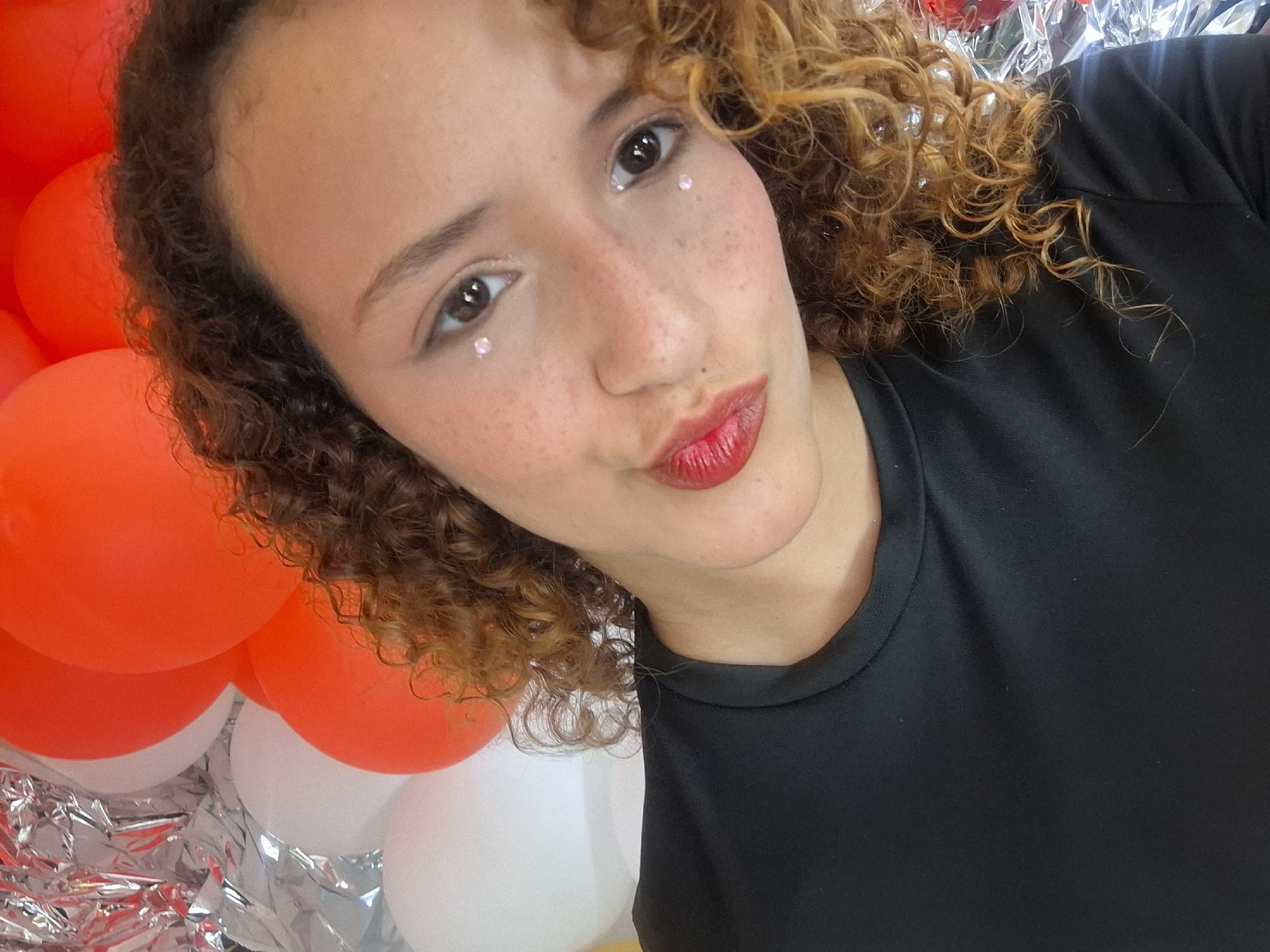 A young woman with curly brown hair and freckles poses close to the camera. She is wearing a black shirt and red lipstick, with small, iridescent decorations under each eye. The background features a mix of orange and white balloons, as well as shiny silver streamers. The location in the city of Playa El Agua, Venezuela is not evident from the image.