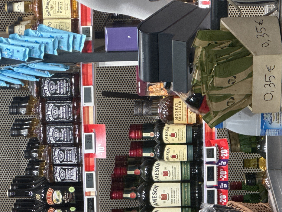 This image depicts the interior of a retail establishment, likely a supermarket or liquor store in Paris, France.
The scene focuses on shelves displaying various alcoholic beverages and a foreground counter area. The background features perforated metal shelving characteristic of commercial retail environments.
On the upper shelf, multiple bottles of Baileys Irish Cream, Jack Daniel's Tennessee Whiskey (in standard and smaller sizes), and Jack Daniel's Tennessee Honey are arranged. Digital price tags show values such as "17," "29," and "27" (implied Euros), along with a prominent red "PROMO" tag indicating a "-10%" discount. Several light-blue wrapped items hang from a vertical fixture, possibly small confectionery or sachets.
The lower shelf presents multiple bottles of Jameson Triple Distilled Irish Whiskey (in various sizes) and a bottle of Monkey Shoulder Blended Malt Scotch Whisky. Digital price tags display "14" (implied Euros) and red "PROMO" tags indicating "-20%" discounts. On a lower level, smaller bottles, including one with yellowish liquid, are visible.
In the foreground, on what appears to be a checkout counter, a black EPSON brand point-of-sale terminal or printer is positioned. Adjacent to it, a light-colored wooden box contains numerous small, light-green and beige pouches, each clearly marked with a handwritten price of "0,35 €." A white plastic bottle with a blue label, identifying it as "lactel Matin léger," is partially visible next to the wooden box. On the far right, a partial view of a white sleeve or garment suggests the presence of a person at the counter. No direct actions or interactions are visible.