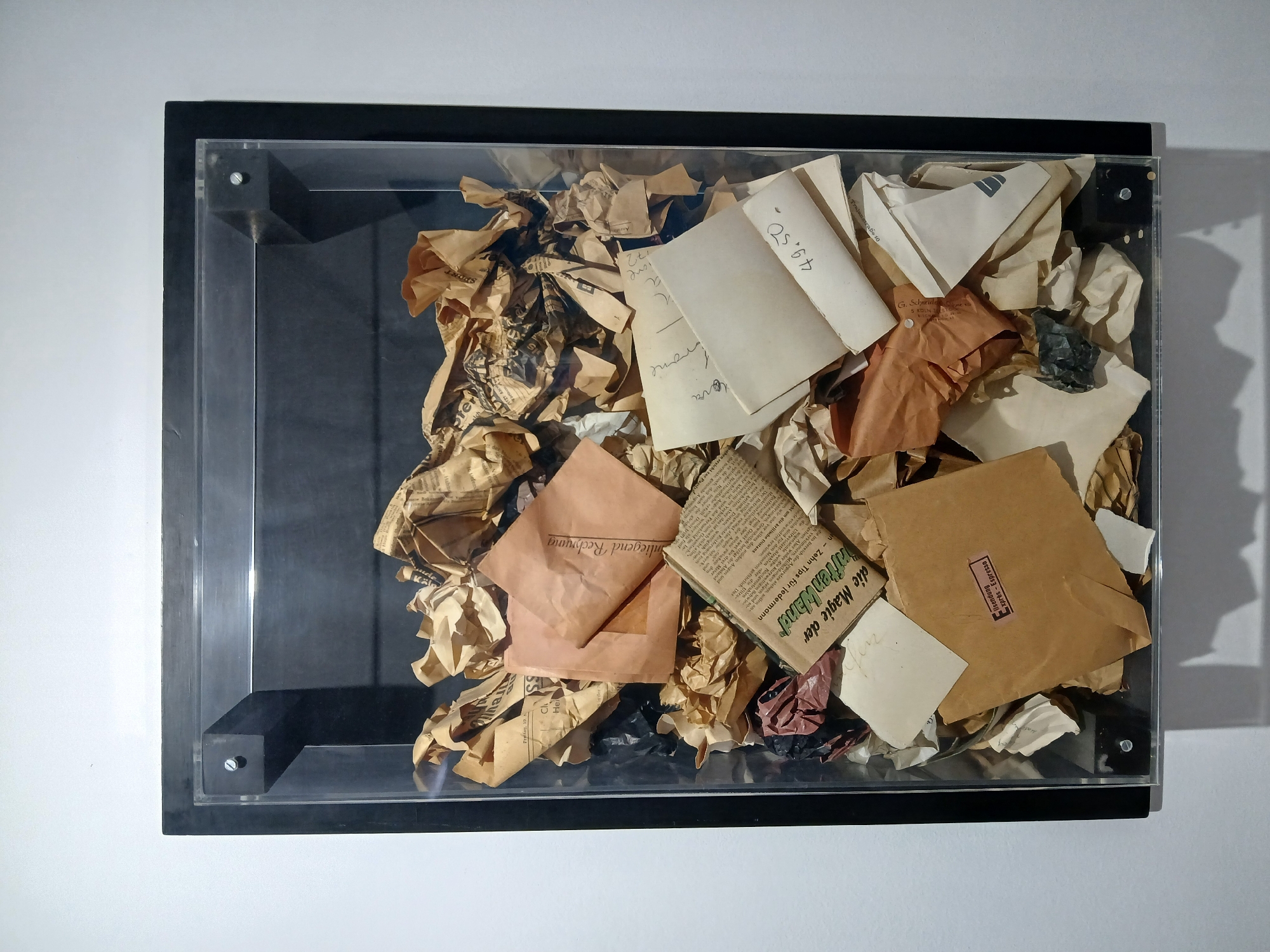The image shows an art installation, a collection of crumpled papers and ephemera enclosed in a clear, rectangular box with a black frame. The papers vary in color and texture, with some appearing aged and yellowed, while others are in brighter shades of brown and white. Text is visible on some of the papers, written in a European language and what appears to be a handwritten numeral and currency symbol. The contents are arranged somewhat haphazardly, filling the box to varying depths. The box is mounted on a white wall, suggesting a gallery or museum setting.