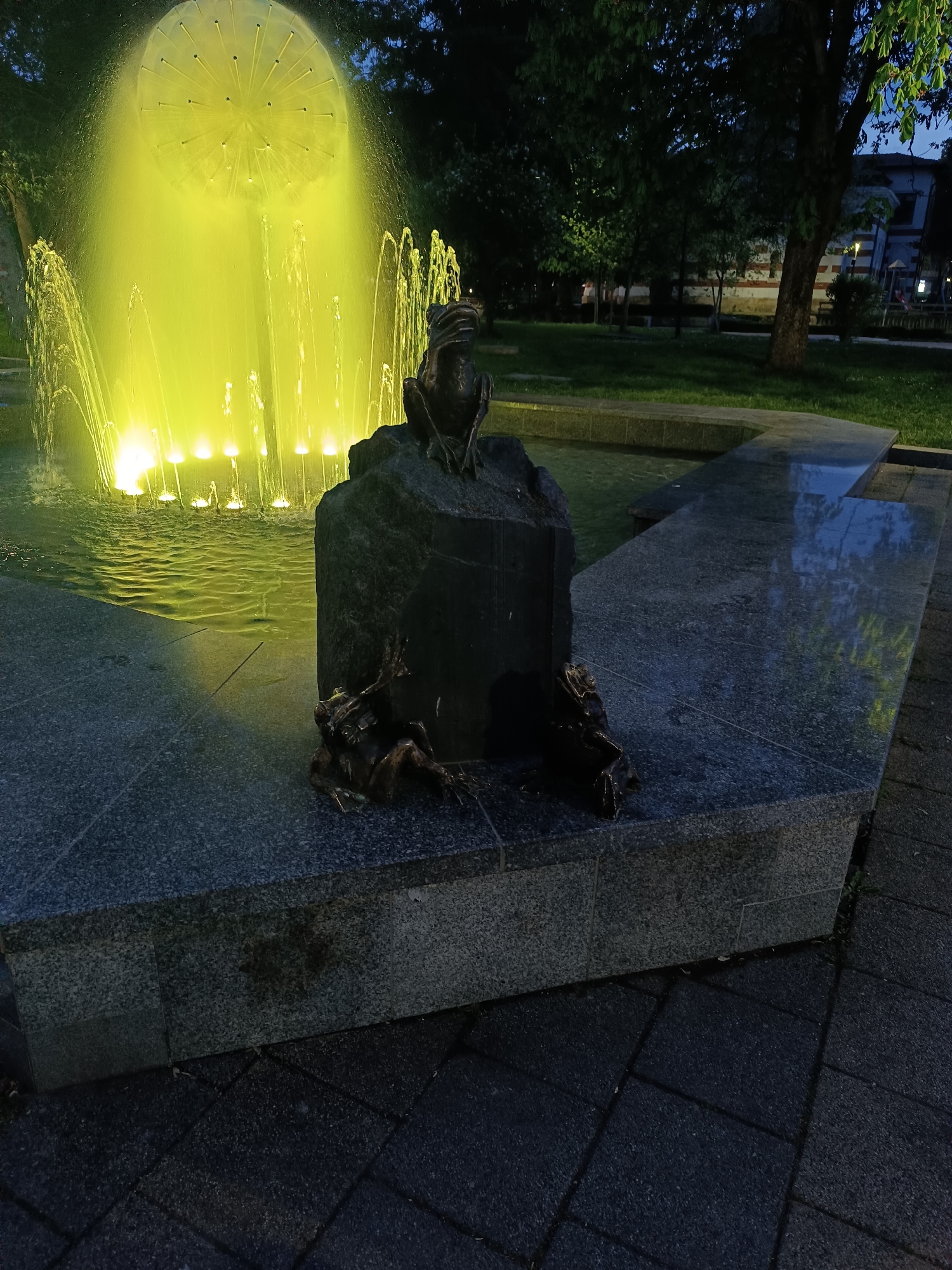 An eye-level shot captures a vibrant night scene featuring a fountain with bright yellow illumination, surrounded by dark granite structures and bronze frog statues. The fountain dominates the left side of the frame, showcasing a central, large spherical element spraying water upwards, and multiple vertical water jets along its perimeter, all bathed in intense yellow light that reflects off the water's surface.
In the foreground, to the right of the fountain, stands a dark, rough-hewn stone monument. Perched atop this stone is a bronze statue of a frog, appearing to gaze towards the illuminated fountain. At the base of the stone, two smaller bronze frog statues are positioned, one on either side, facing towards the viewer and the fountain respectively.
The immediate ground around the fountain and monument consists of dark, polished granite tiles, some of which reflect the yellow light. Beyond the fountain and the granite platform, the scene recedes into a dark park-like area with numerous trees and foliage, dimly lit by the distant ambient light. A building with some visible architectural features can be faintly seen in the far background through the trees. The overall time of day is nighttime, indicated by the dark sky and reliance on artificial lighting. No text is visible in the image.