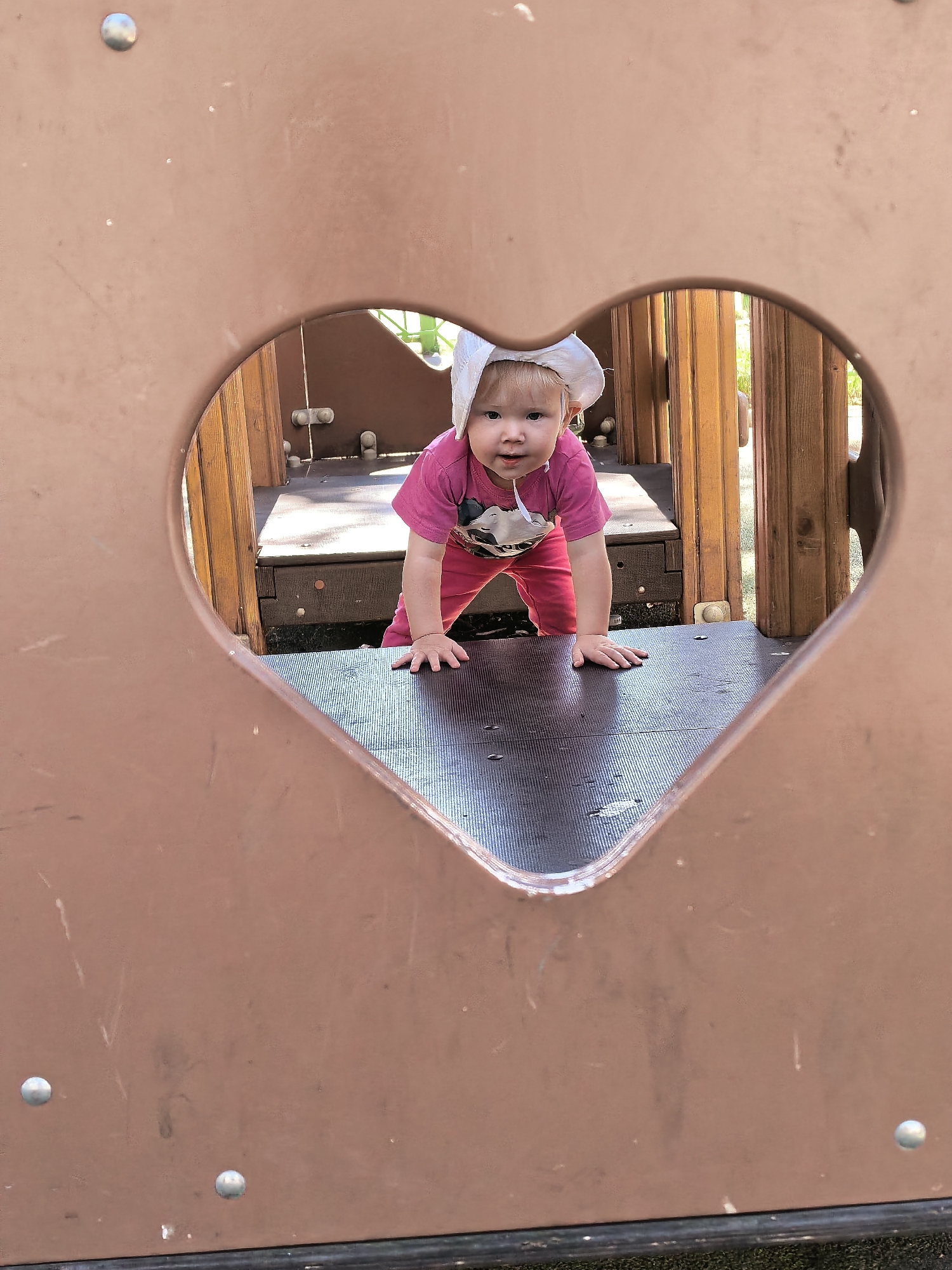 A young child wearing a pink shirt, pink pants, and a white hat is crawling on a playground structure, looking at the camera. The child is viewed through a heart-shaped opening in a brown panel of the playground. The child appears to be a toddler, with blonde hair and light skin. The playground structure is made of wood and brown plastic. The image appears to have been taken outdoors during the daytime. There is no information in the image that would indicate a specific location in the city of Sterlitamak, Russia.