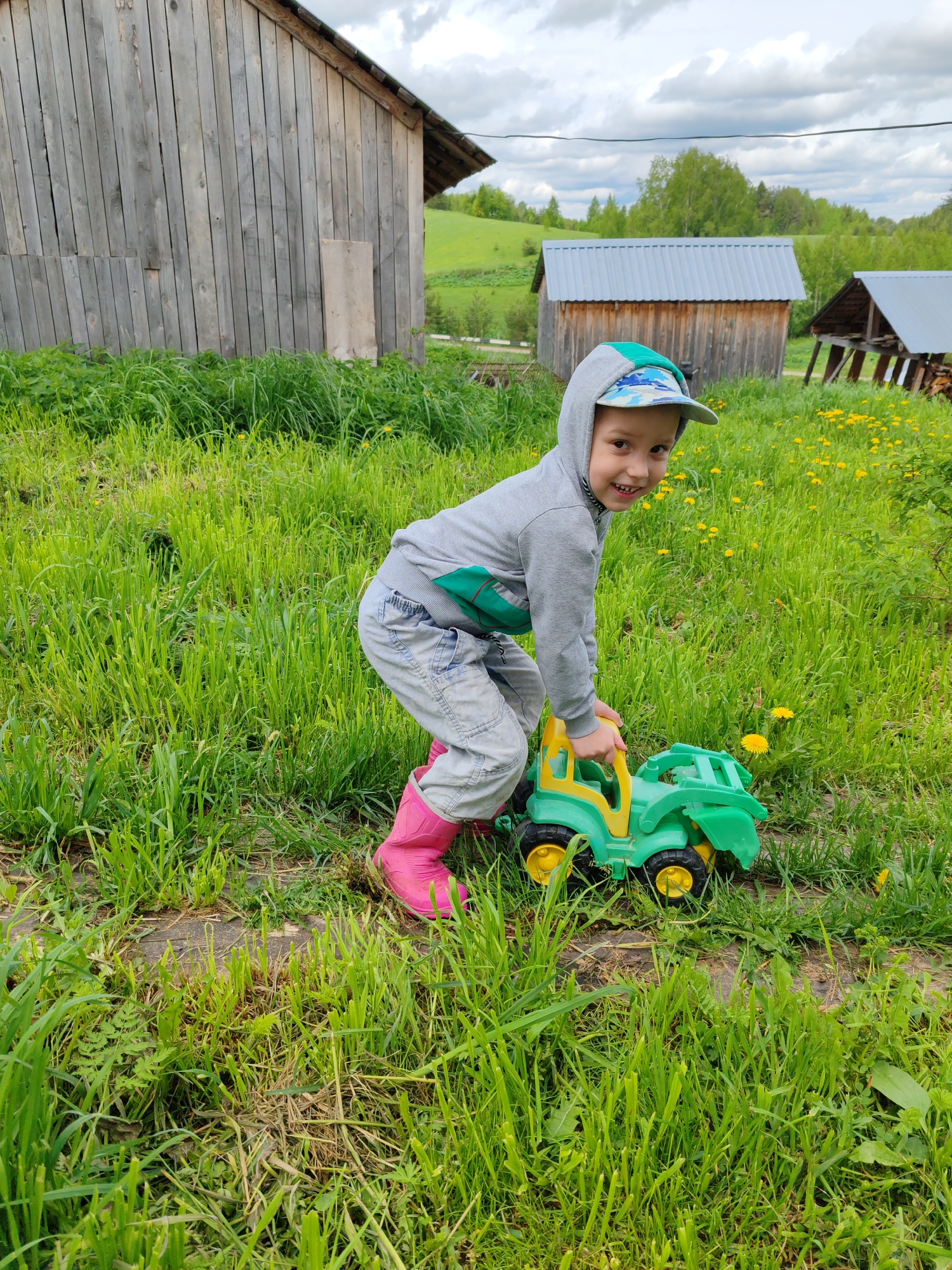 A young boy plays with a green and yellow toy tractor in a grassy field near Чухлэм, Russia. He is wearing a grey hooded sweatshirt, light blue pants, a baseball cap, and bright pink boots. He is bent over the tractor, looking towards the viewer with a smile. The field is full of tall green grass and some yellow wildflowers. In the background, there are several wooden buildings, including a large weathered barn to the left and two smaller structures with metal roofs to the right. A rolling, green hillside is visible behind the buildings. The sky is overcast with grey clouds.