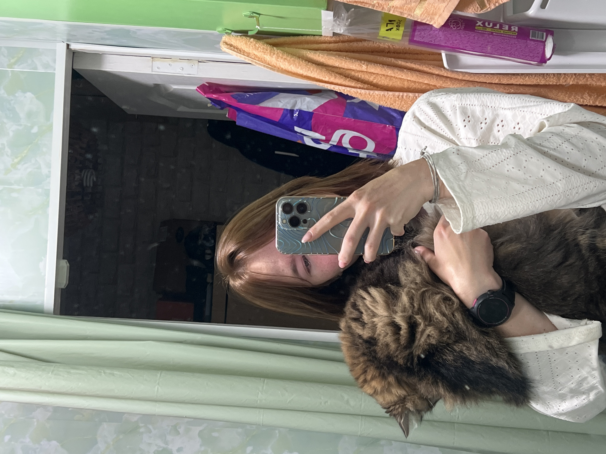 The image, rotated 90 degrees clockwise, captures a mirror selfie taken by a person holding a large cat in an indoor setting in Chaikovsky, Russia.
The central subject is a person, likely female, with long brown hair, whose face is partially obscured by a smartphone. Her left eye, nose, and part of her cheek are visible. She is wearing a white long-sleeved top with eyelet embroidery. A silver bracelet adorns her left wrist, and a black smartwatch with a pink inner bezel is worn on her right wrist. She holds a large, fluffy, brown and black tabby cat close to her chest, with its head nestled against her and its dense fur prominent.
The person is holding a silver-blue patterned smartphone, likely an iPhone with a triple camera system, to capture the reflection.
The background elements are visible both in the mirror's reflection and above it. The mirror reflects a dimly lit, dark interior with indistinct shapes, possibly furniture. Above the mirror, there is a light green shelf or cabinet top. On this surface, an orange folded towel is visible, featuring a yellow sticker with the reversed Cyrillic text "70ВК". Next to the towel, a purple plastic bag with large, partially obscured white reversed lettering is present. A white rectangular tray holds a purple package, clearly labeled with the reversed Cyrillic text "РЕЛАКС" (RELAX), along with smaller, illegible Cyrillic script. A light green curtain or fabric is visible in the foreground, partially obscuring the bottom of the mirror. A white door or window frame is also present.
The scene depicts a casual, personal moment of an individual interacting with their pet at home, potentially in the evening given the dimly lit reflection.